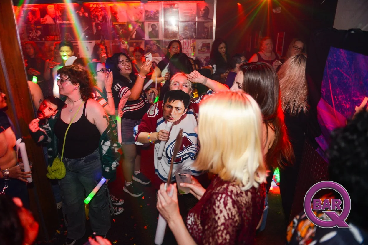 Women and men dancing at a crowded nightclub with colorful lights and posters on the wall, with some people holding glow sticks and drinks.