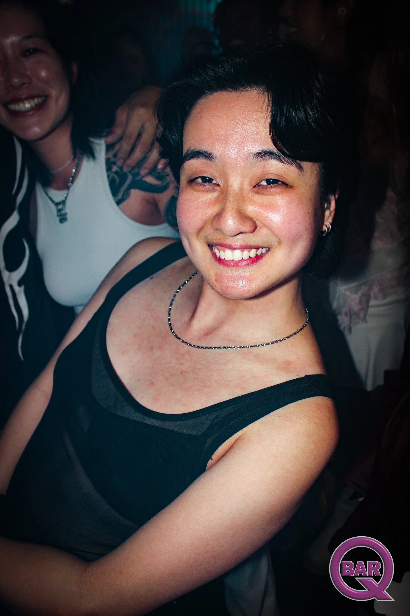 Two women smiling in a dark setting, one wearing a black sleeveless top and the other wearing a white top. One woman has short dark hair and the other has longer hair, and they are close together.