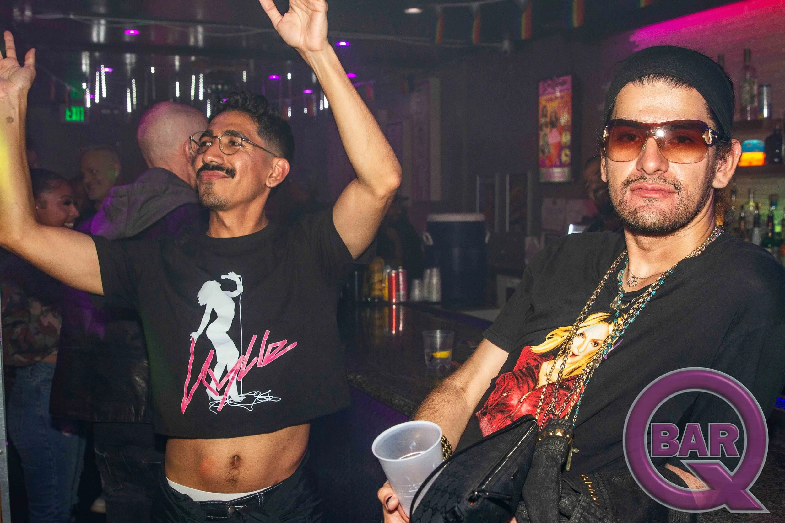 Two men at a nightclub, one in a black t-shirt with pink graphic, the other in a black t-shirt with a graphic of a woman, enjoying the party with drinks in hand.