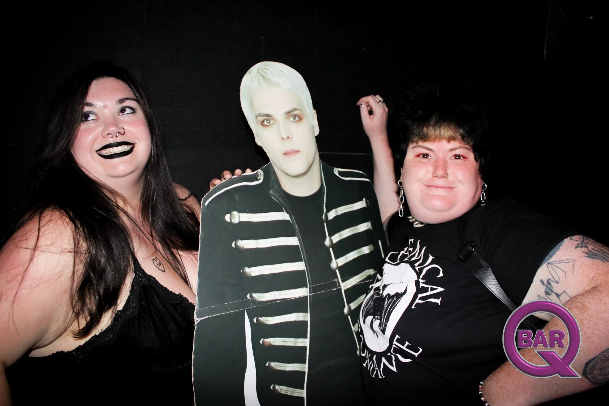 Two women posing with a cardboard cutout of a man with platinum blonde hair and a black and white striped jacket. One woman has dark hair, black lipstick, and a nose ring, and the other has curly dark hair and earrings. They are in a dark setting, po