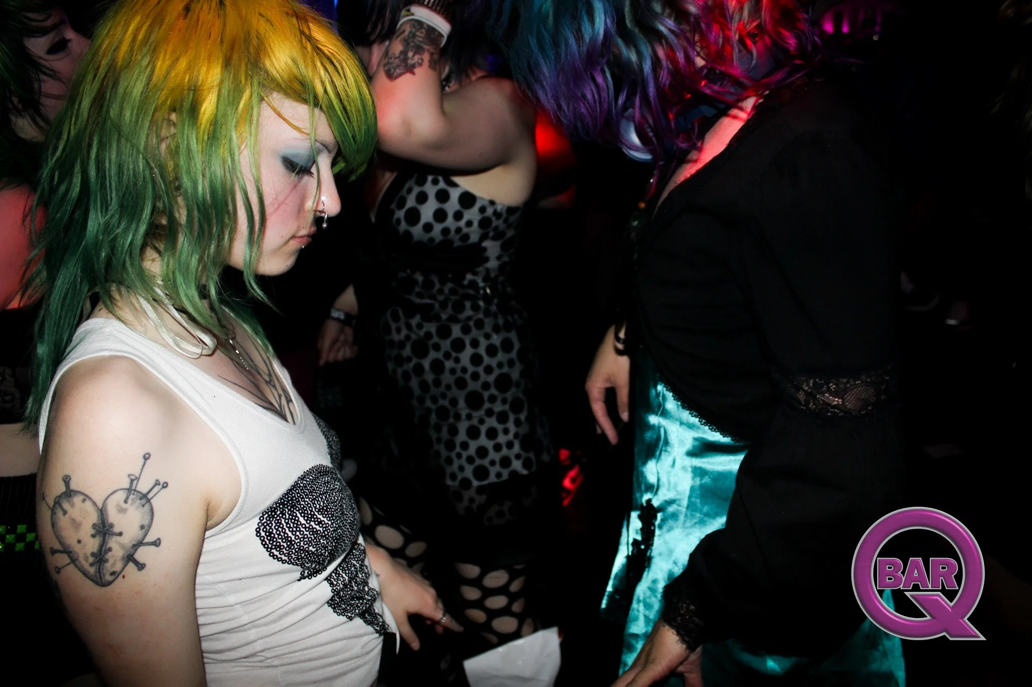 A young woman with rainbow-colored hair and tattoos on her arm, wearing a white tank top with black lace, is in a dimly lit social setting with other women around her, some with colorful hair, and a neon "Bar" sign in the background.