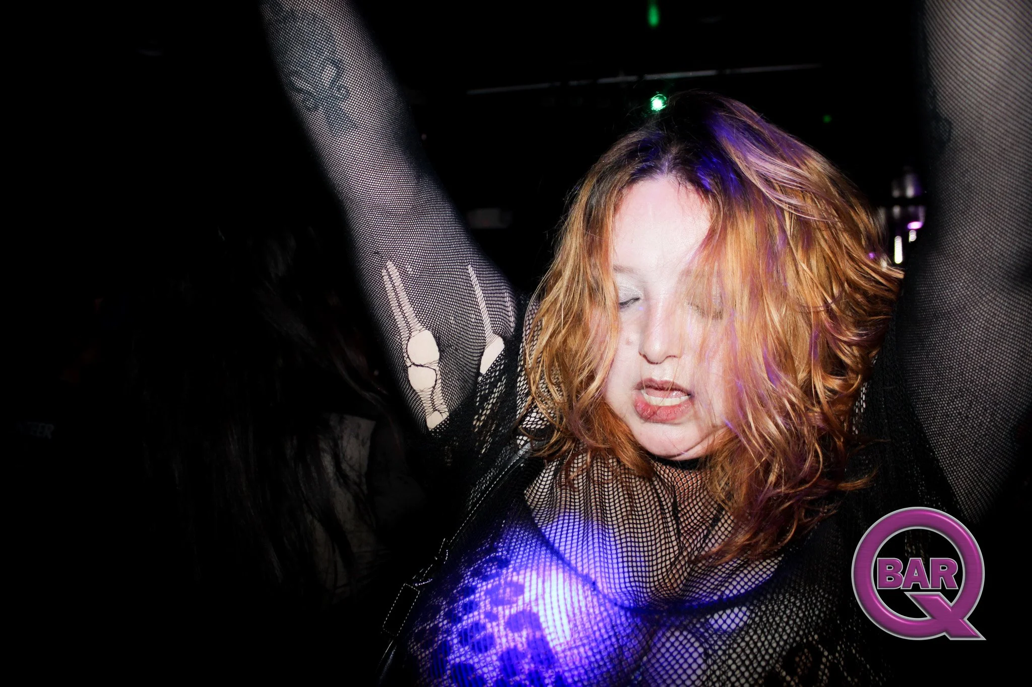 A woman with curly red hair dancing in a nightclub with black fishnet clothing and colorful lights.