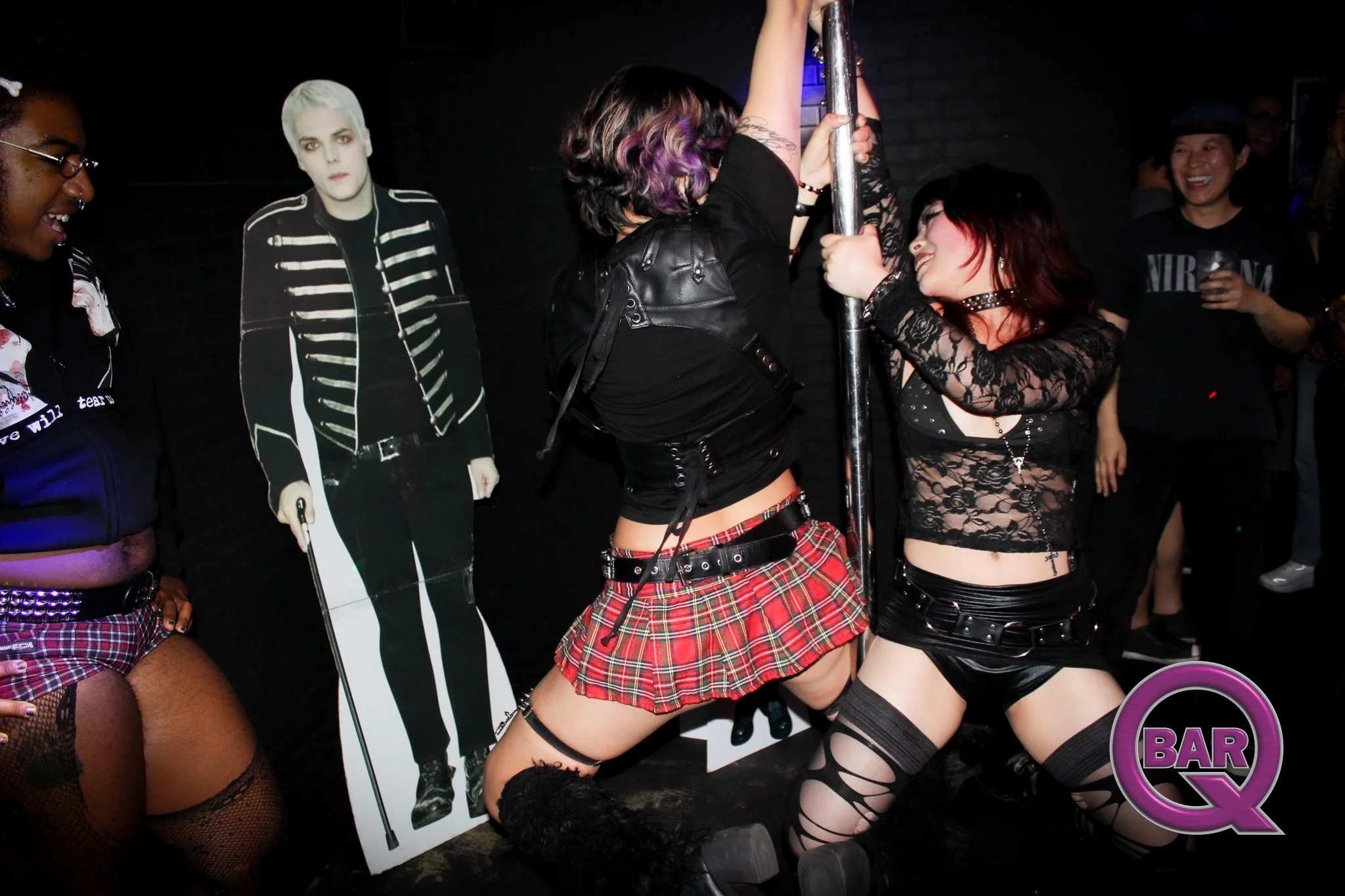 People dancing and socializing at a bar, with a cardboard cutout of a person dressed in a black and white outfit in the background, and a neon sign that reads 'BAR' in the lower right corner.