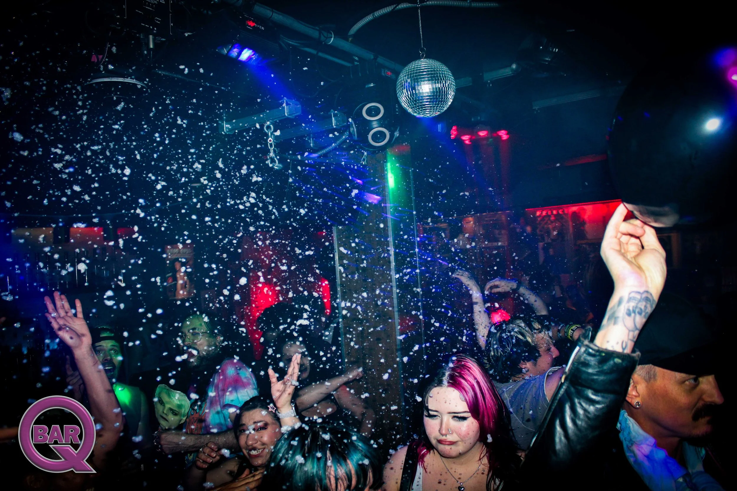 People dancing and enjoying a party inside a nightclub with a disco ball and colorful lights, with foam or snow falling from above.