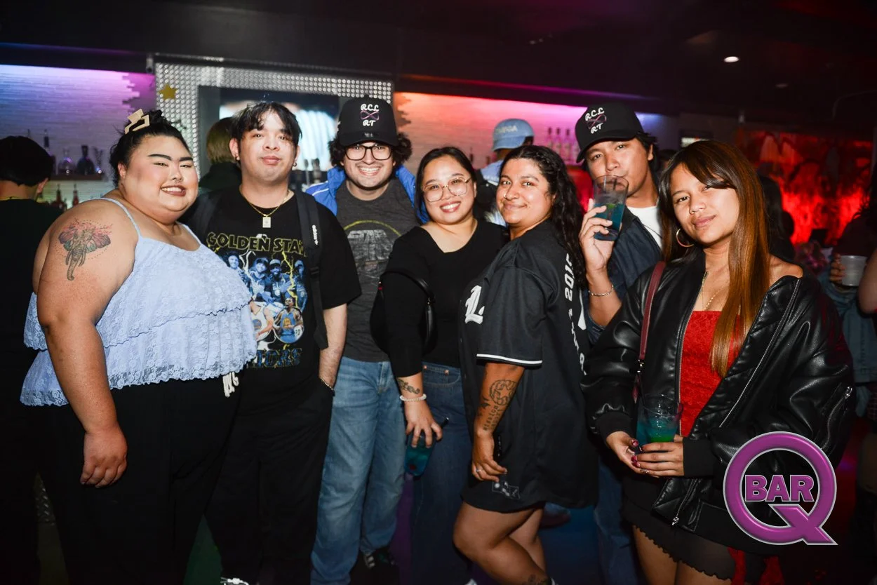 A group of seven diverse women and men posing in a bar with colorful neon lights, some holding drinks, smiling at the camera.