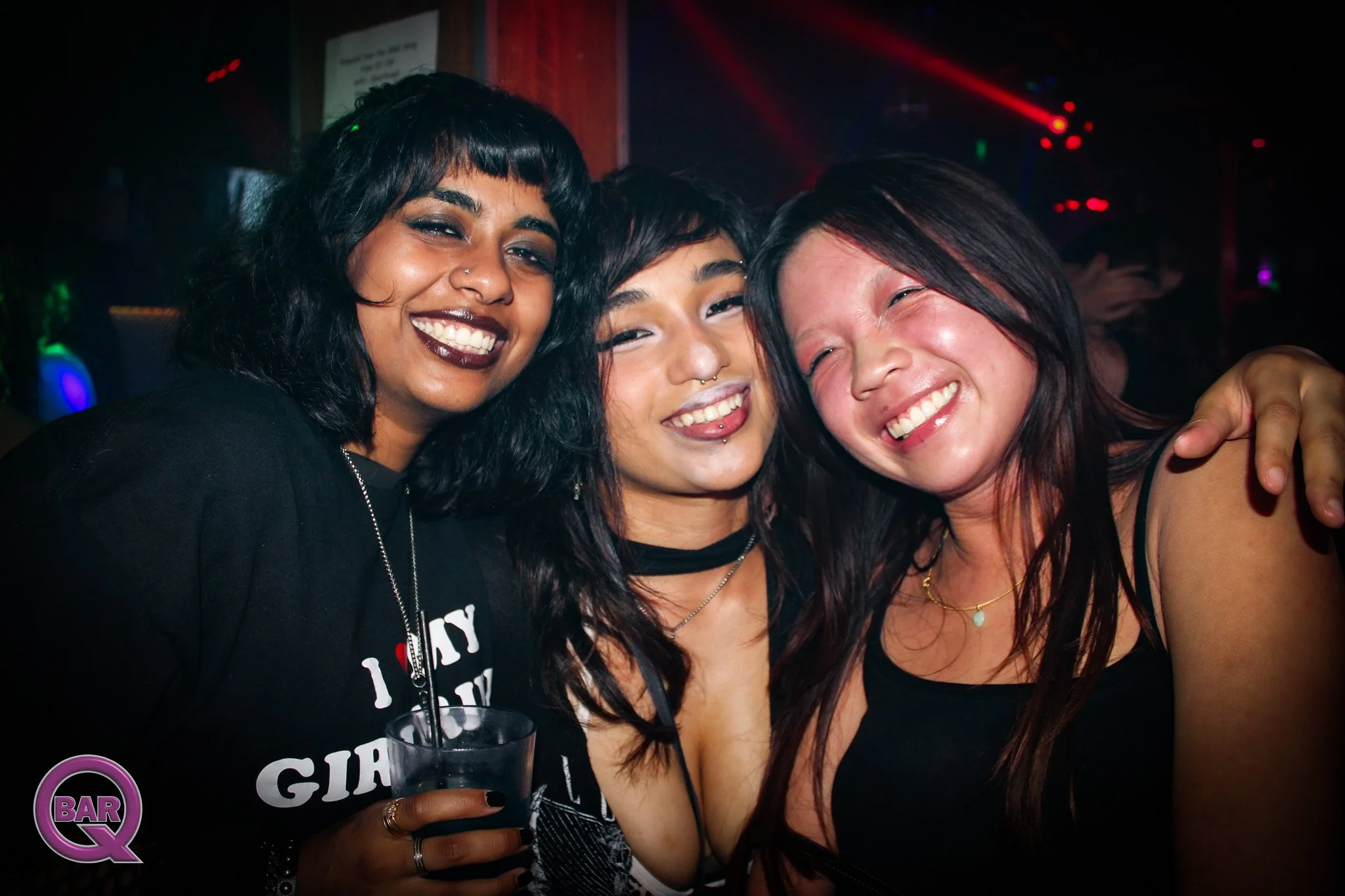 Three women smiling, posing closely together in a dance club with colorful lights in the background, one holding a drink.