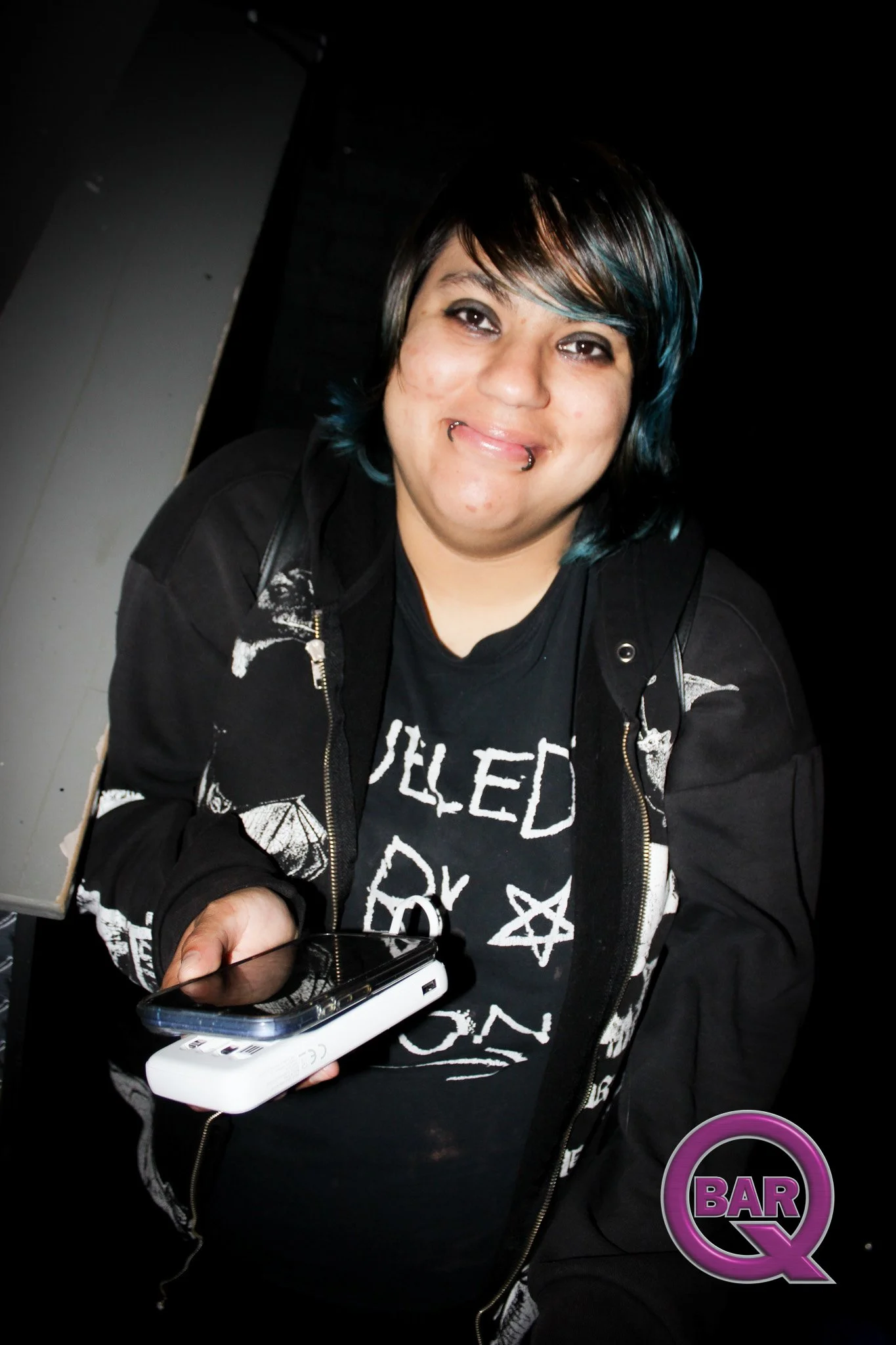 A person with short dark hair with blue streaks, smiling, holding a smartphone, wearing a black Stone Led Play T-shirt and a black jacket, in a bar or club environment.