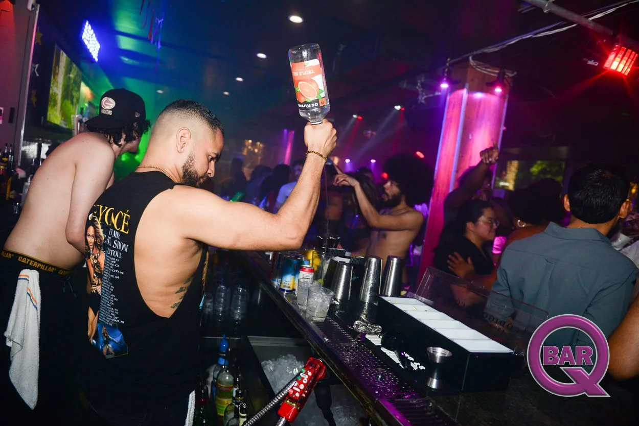 A crowded nightclub with people dancing and socializing. A bartender is pouring a mixed drink from a bottle into a glass, while others dance in the background. The bar area is busy with bottles and glasses, and colorful lights illuminate the scene.
