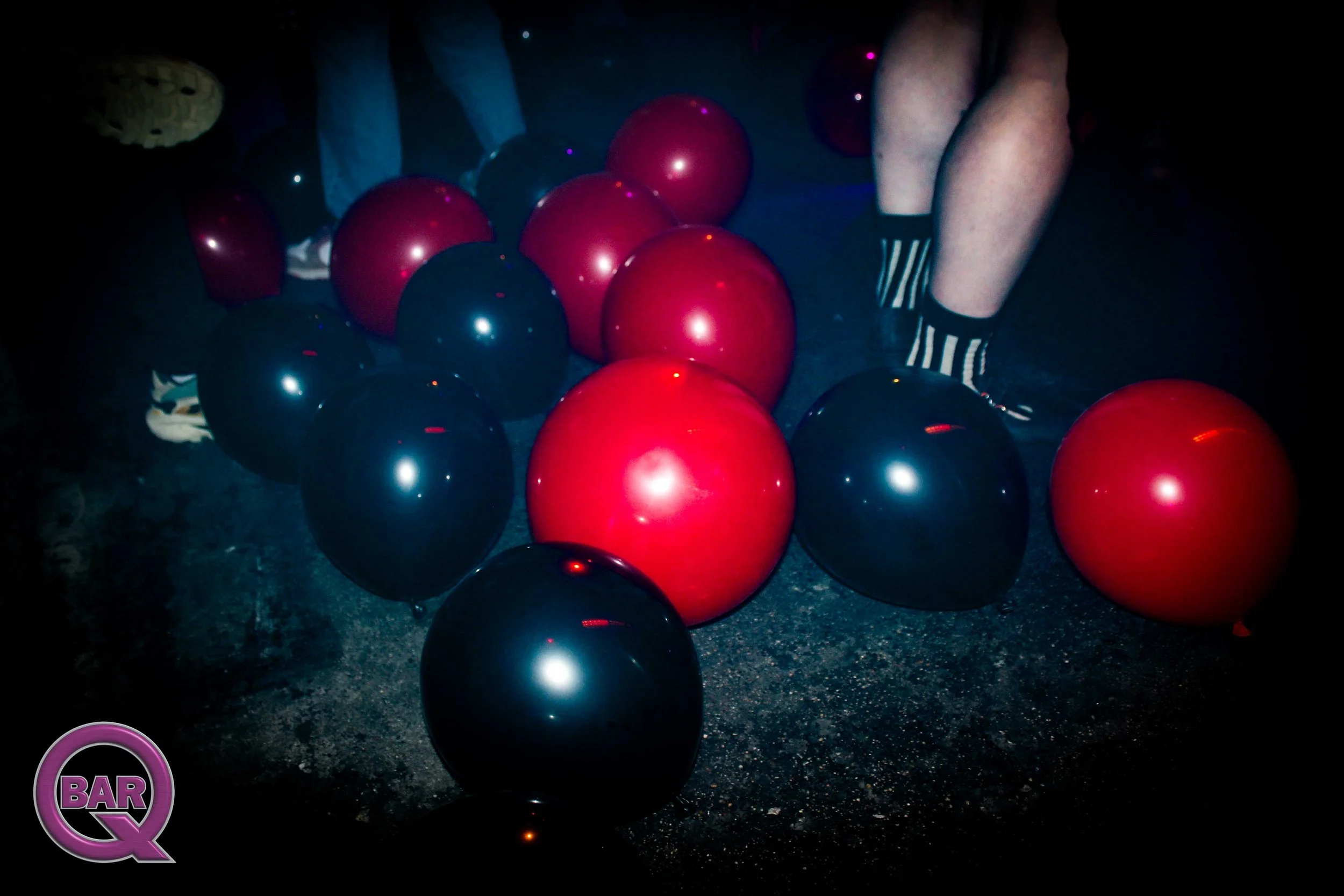 Red and black balloons scattered on the dark floor with people's legs in striped socks visible.