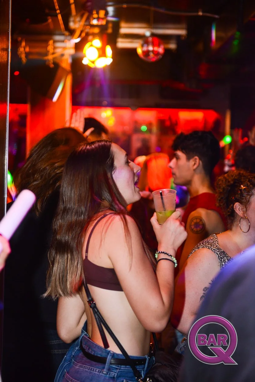 A woman in a black bra top and jeans holding a drink and dancing at a nightclub with colorful lights, surrounded by other people enjoying the music and socializing.
