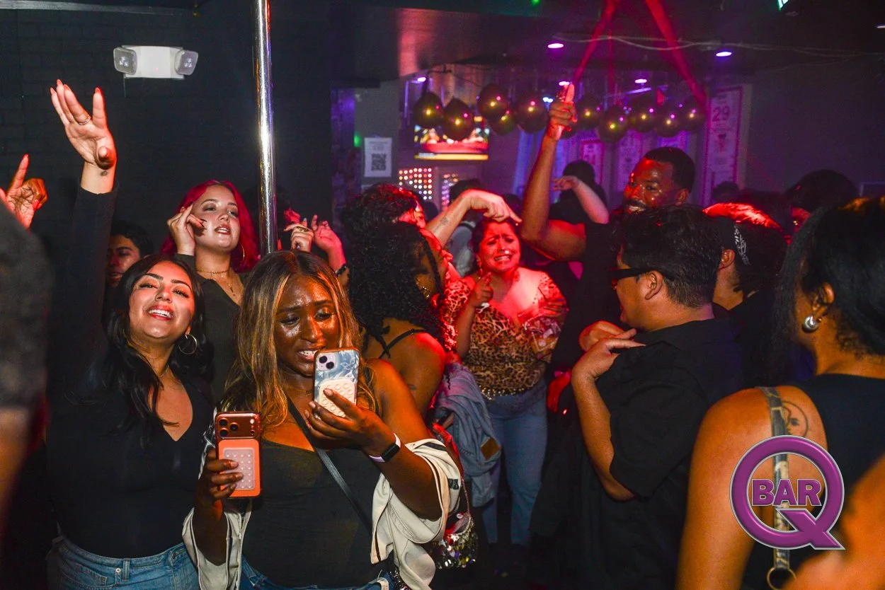 Group of people dancing and enjoying at a nightclub with colorful lights and balloons, some holding phones and taking pictures.