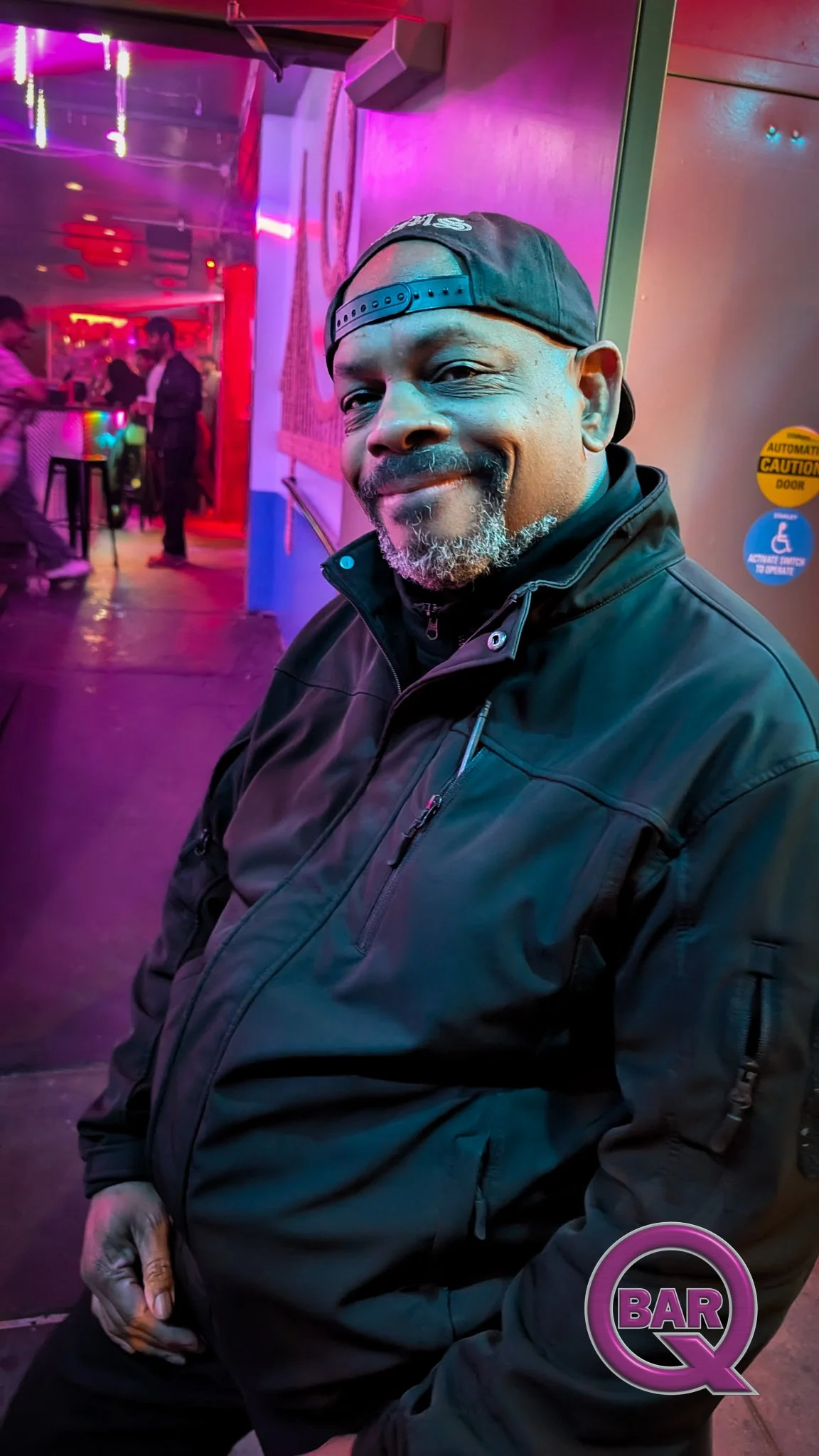 A man with a beard, wearing a black baseball cap backwards and a black jacket, sitting in a bar or nightclub with colorful neon lights and other people in the background.