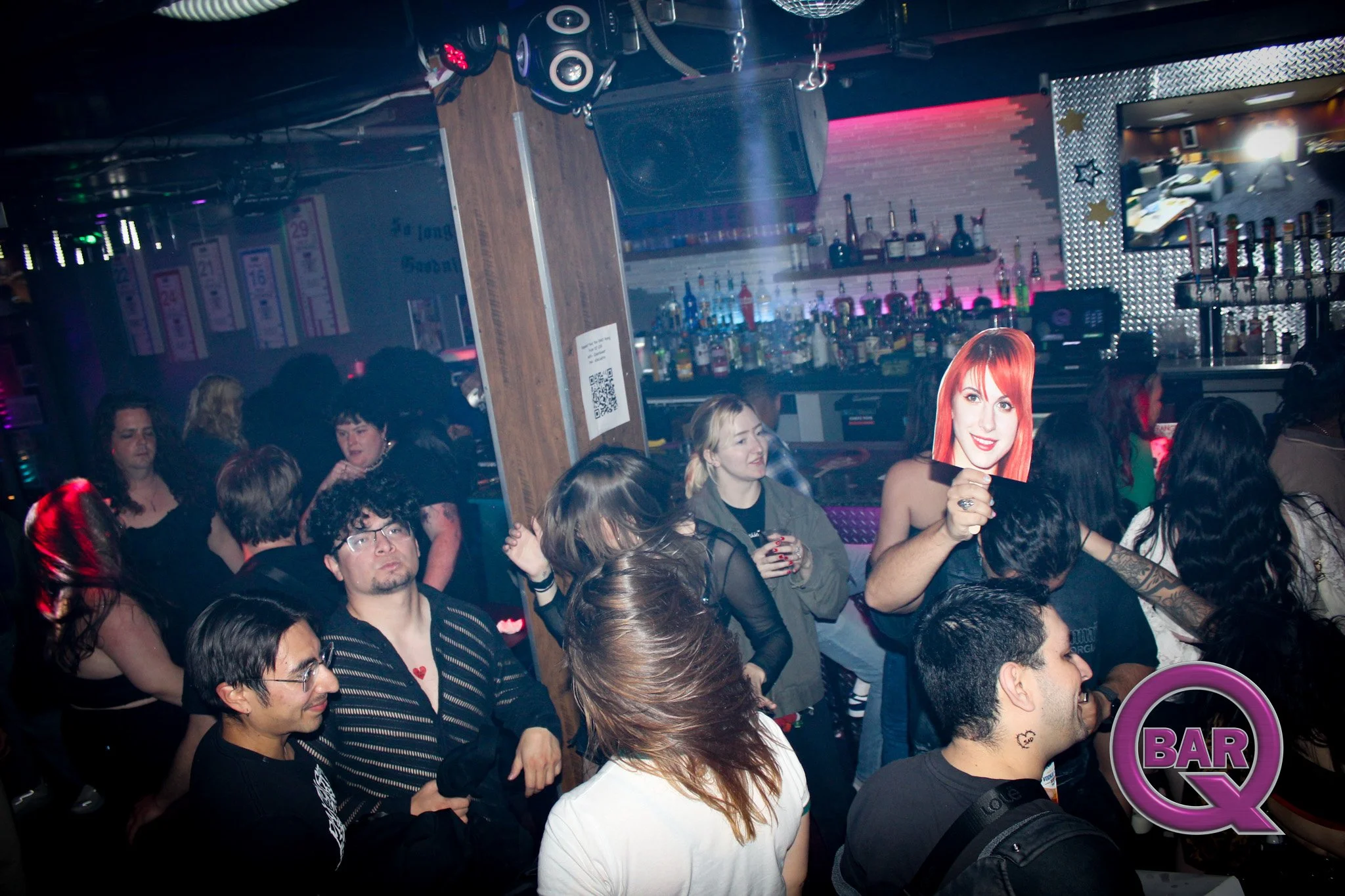 Crowd of people at a bar or club, some holding drinks, with a bar area in the background illuminated with pink and purple lights, and various bottles on shelves. A woman is holding a cutout of a woman's face near her own face. The environment appears