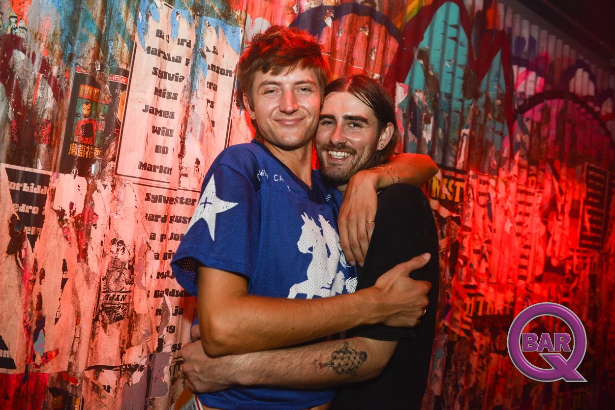 Two men hugging and smiling at a bar or club with colorful, graffiti-like wall art in the background, one wearing a blue sports jersey and the other in a black shirt, with a purple round logo labeled 'BAR Q' in the corner.