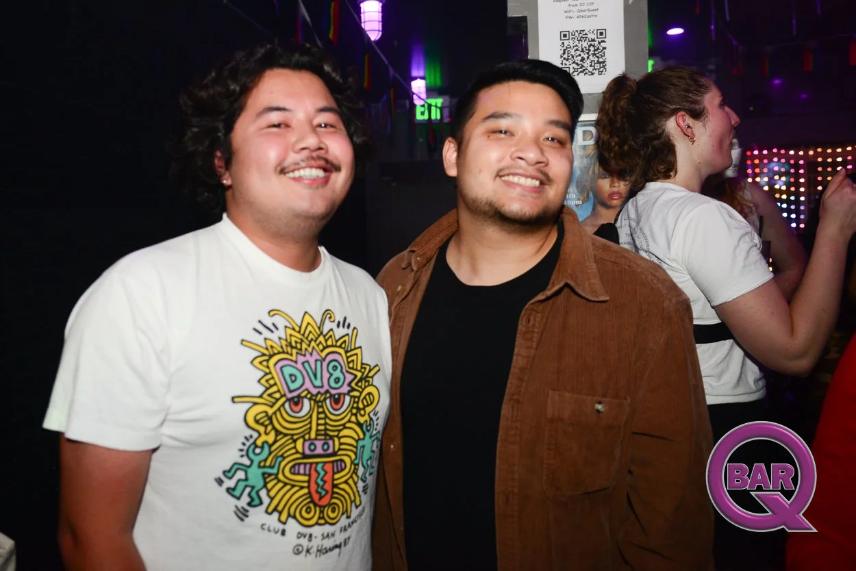 Two smiling men posing together in a bar or nightclub with colorful lights and people in the background, one wearing a white t-shirt with a graphic design and the other in a brown jacket.