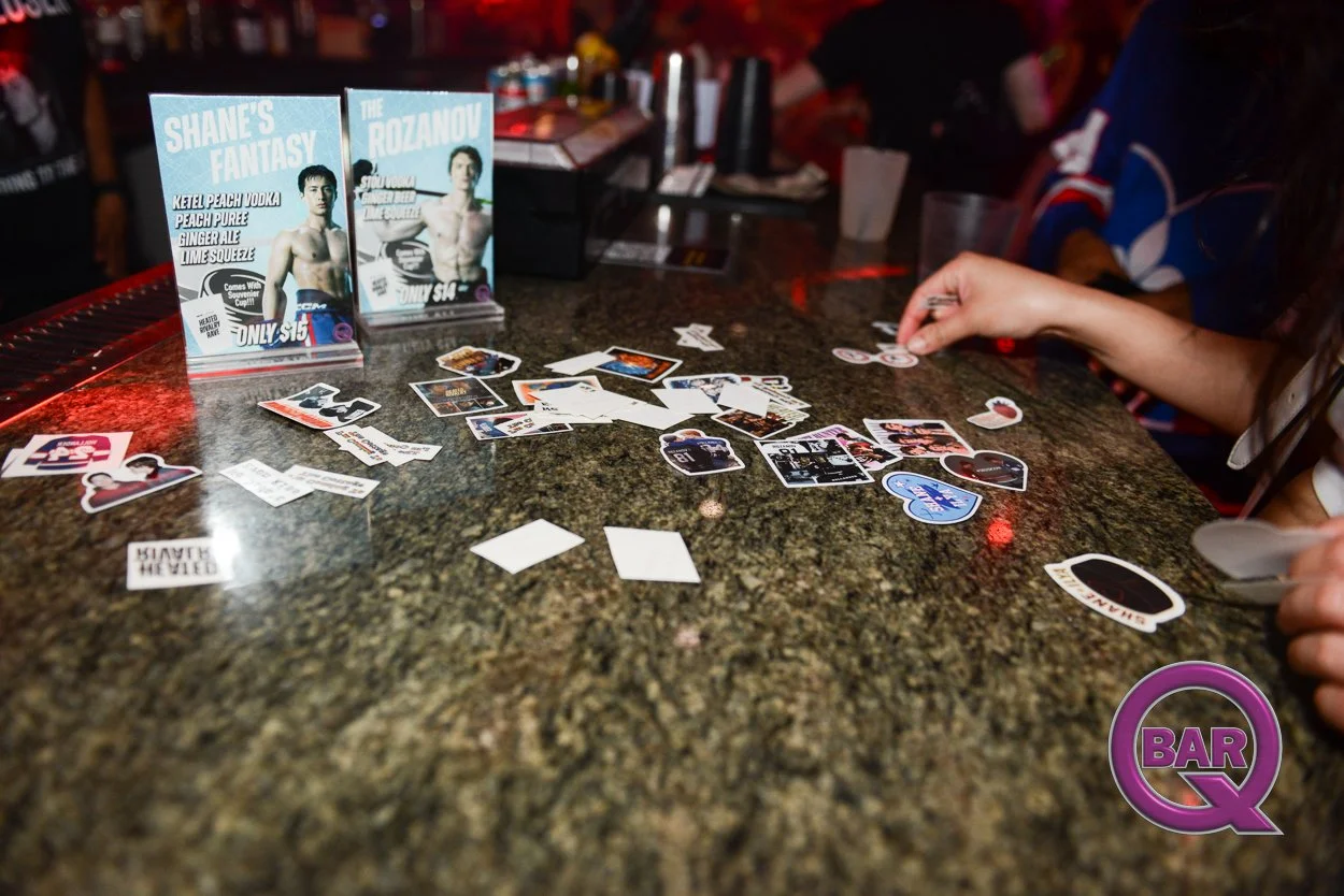 A bar scene with a dark granite countertop covered with scattered stickers, playing cards, and promotional flyers for Shane's Fantasy and The Rozanov. A person's hand is reaching towards the stickers, and the background shows blurred bar equipment an