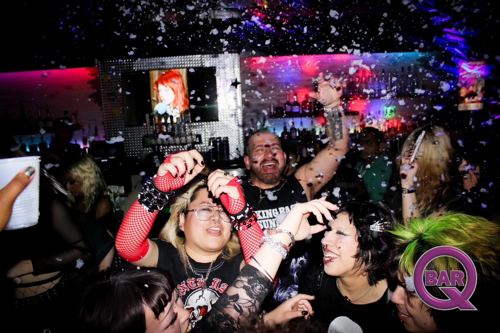Crowded nightclub scene with people dancing and celebrating, colored lights, and a large screen displaying the image of a red-haired woman.