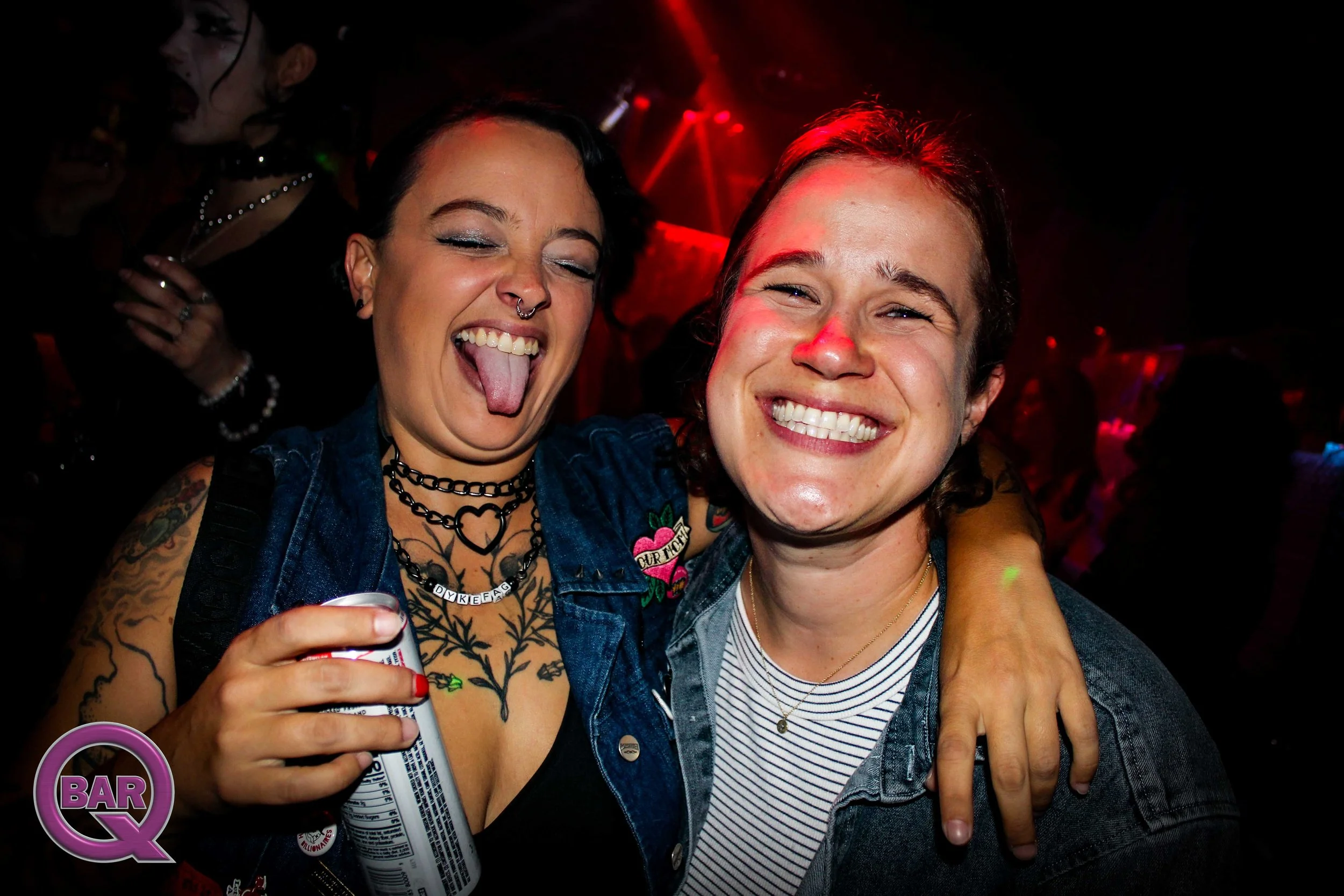 Two friends smiling and hugging at a nightclub or bar, with one holding a can of soda. The woman on the left has dark hair, tattoos, a septum piercing, and wearing a denim jacket and necklaces. The man on the right has curly hair, a broad smile, and 