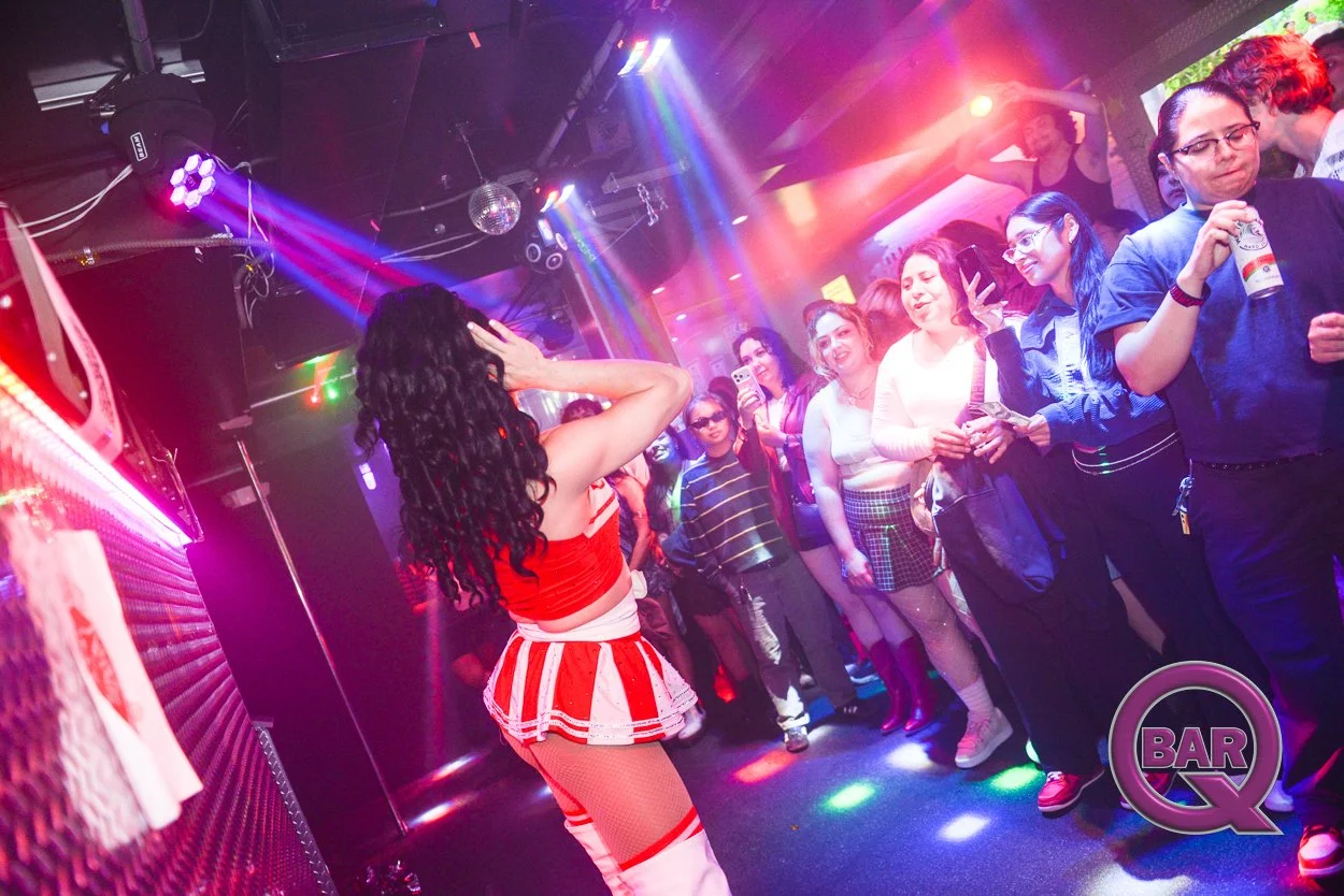 A performer in a red and white striped outfit with long black curly hair is on stage, singing or dancing in a club with a crowd of women watching and taking photos. The club has colorful lighting, a disco ball, and a bar logo visible.