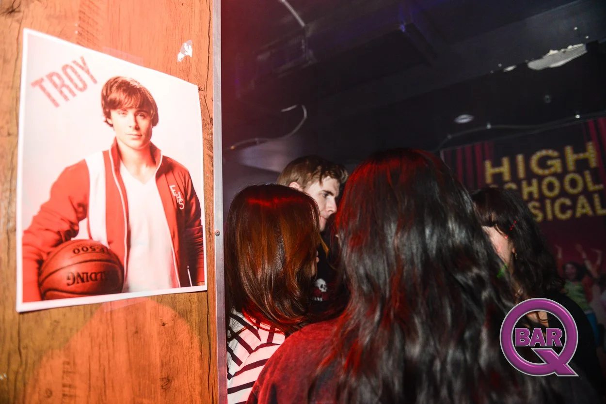 Group of people at a bar or nightclub with a poster of Troy, a young man holding a basketball, on the wall. The background has a sign that says 'High School Musical'.