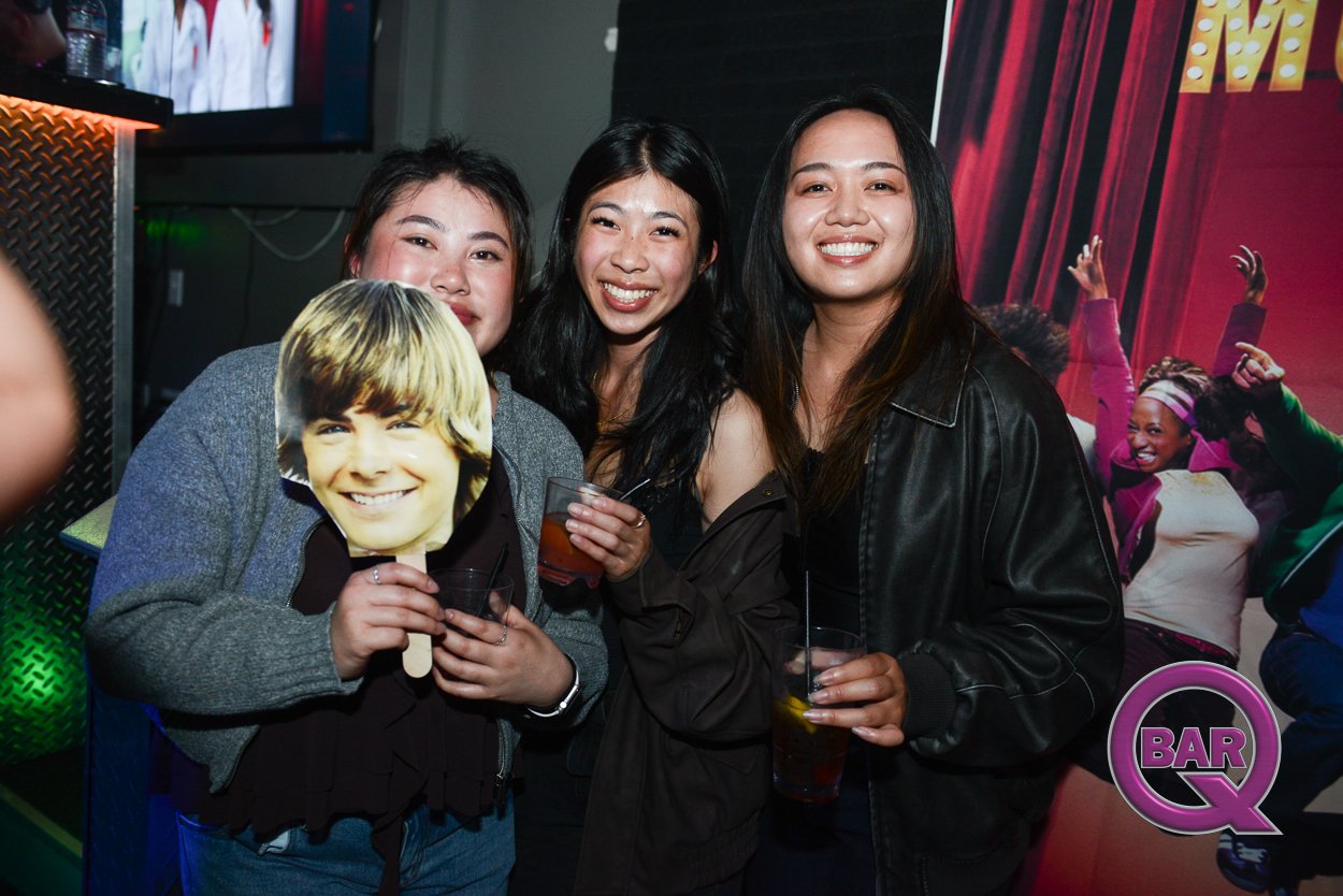 Three women at a bar holding drinks and a mask of a young boy with blond hair. The women are smiling and standing close together. The background features a poster with people dancing and a bar logo.