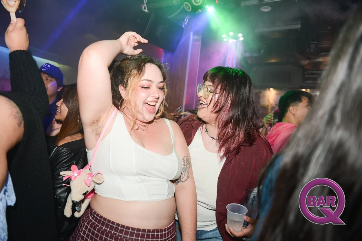 Two women are dancing and laughing in a crowded bar or club with colorful lights. One woman has curly hair, tattoos, and is wearing a white crop top, while the other woman has straight hair, glasses, and is wearing a red jacket.