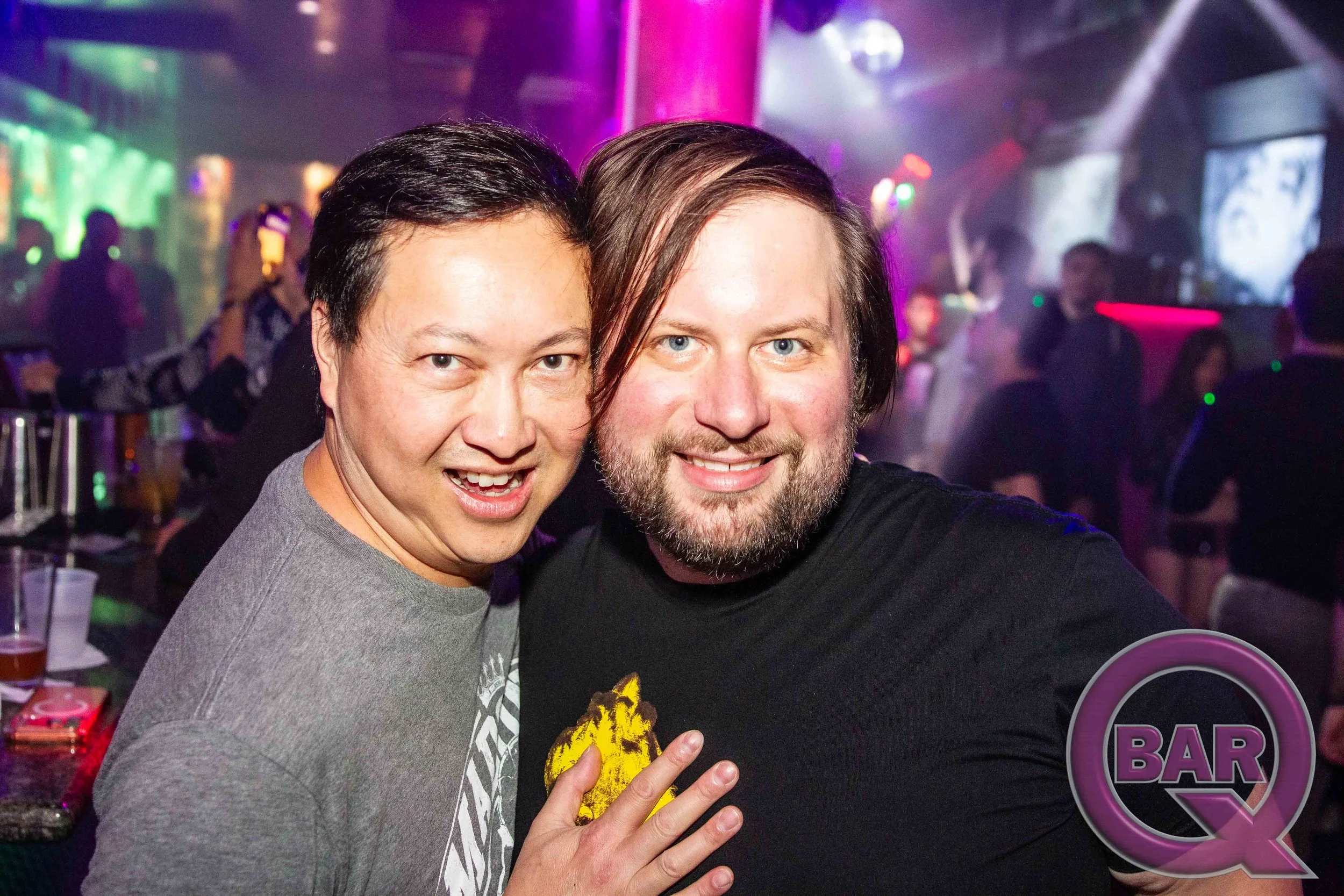 Two men smiling and posing for a photo in a bar or club, with colorful lights and blurred people in the background.