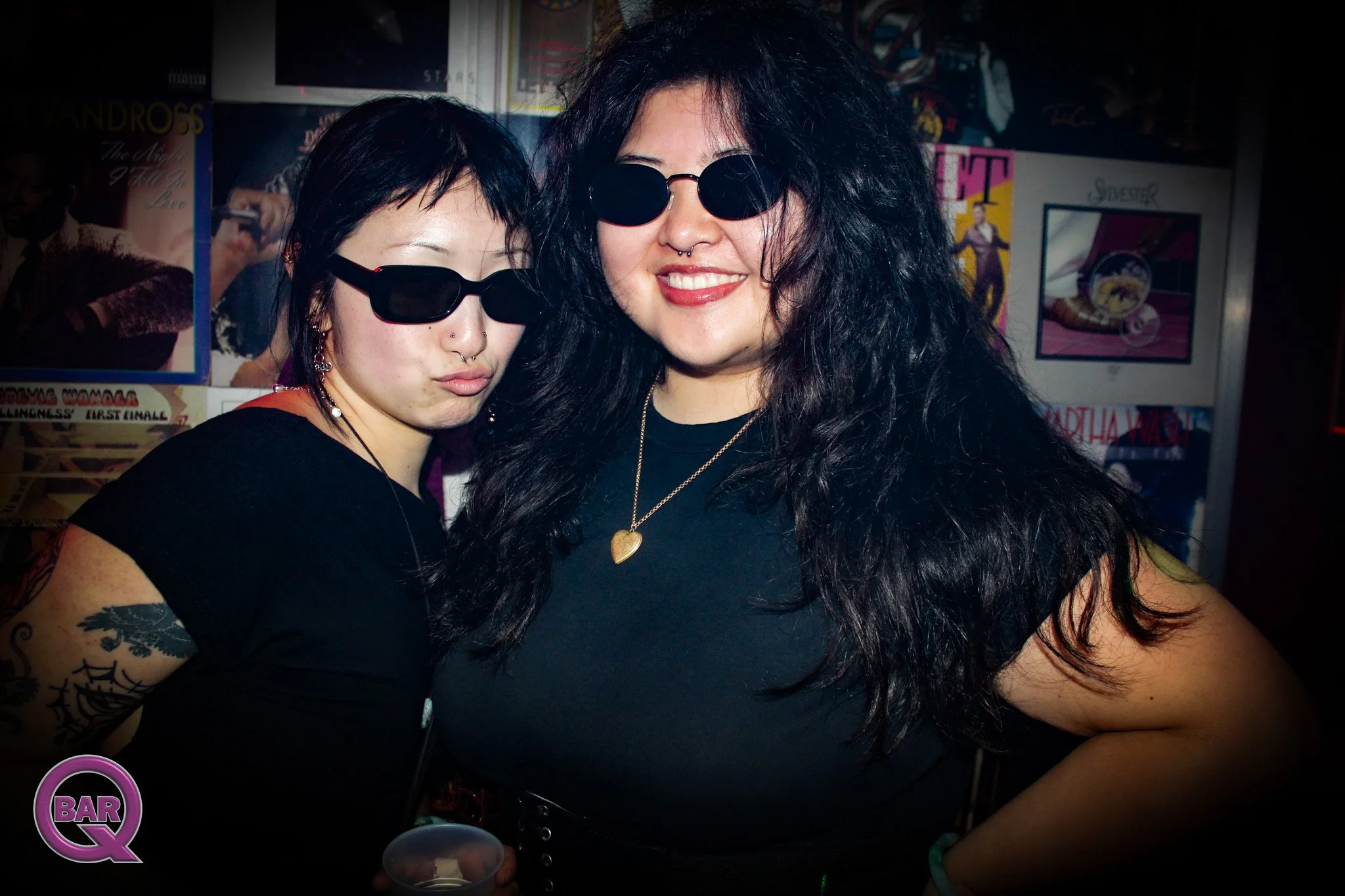 Two women with dark curly hair and sunglasses pose together in a dark indoor setting, smiling at the camera. One woman has glasses and a septum piercing, and the other has tattoos on her arm and wears a heart pendant necklace.