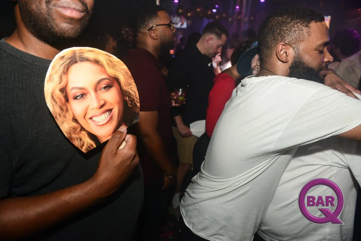 Group of people dancing at a nightclub with purple lighting. A man in the foreground is holding a paper cutout of a smiling woman with blonde hair. There is a purple and pink "Bar Q" logo in the bottom right corner.