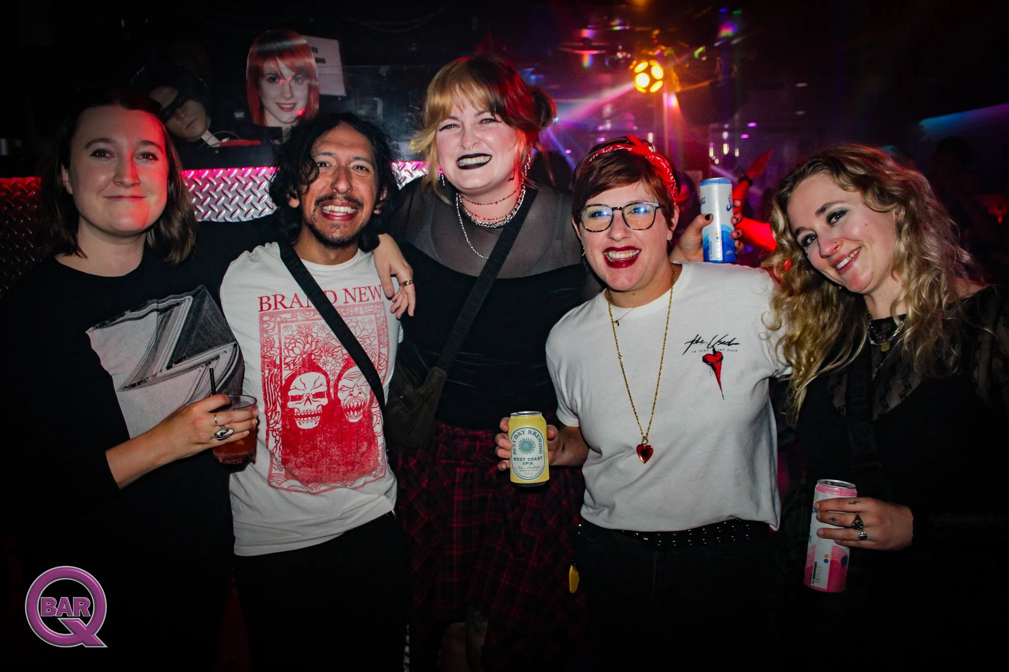 Group of five people smiling and posing together at a bar or nightclub, with colorful party lighting and a DJ in the background.