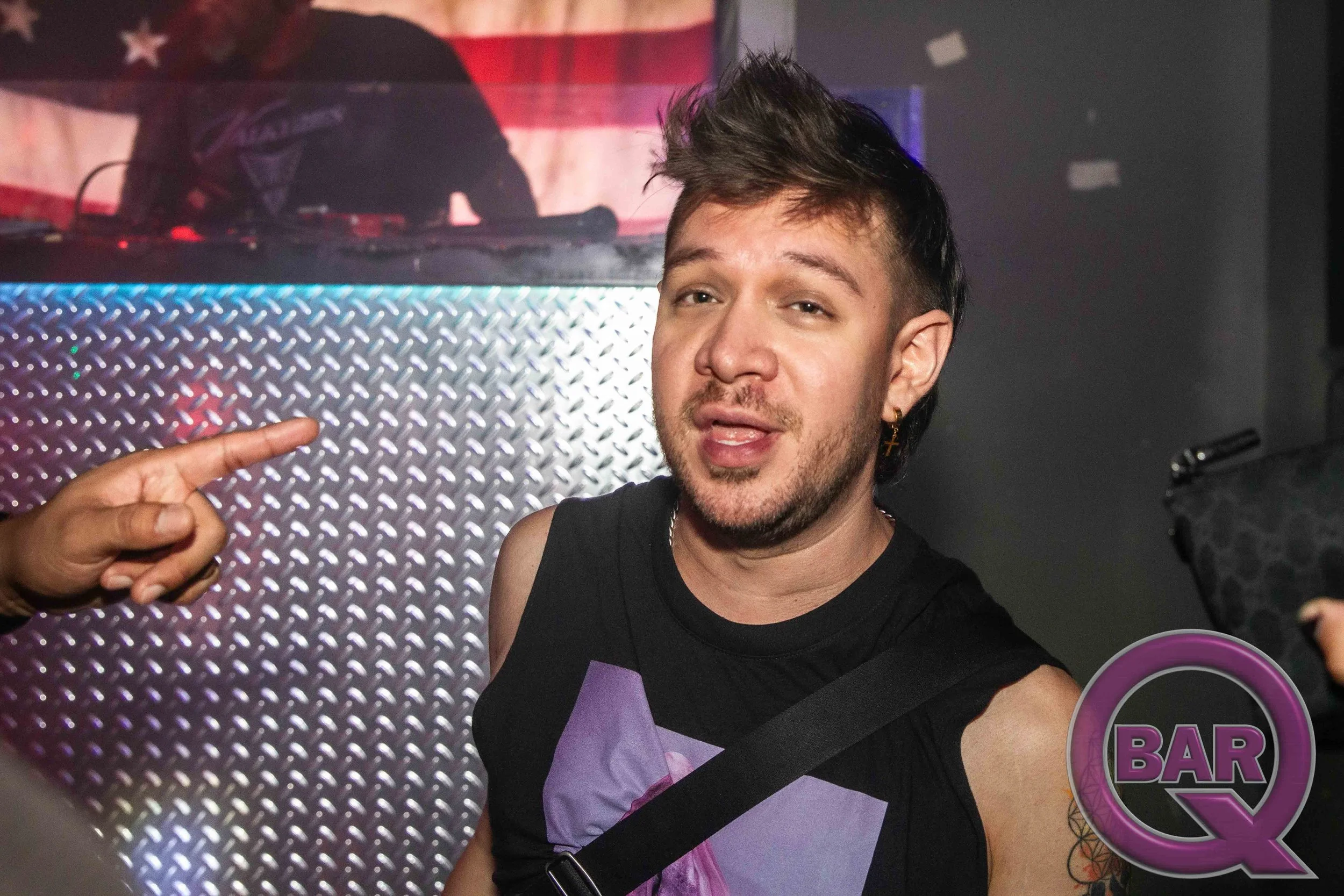 Young man with spiked hair taking a selfie at a bar, wearing a black tank top with purple geometric design and a sash with purple accents.