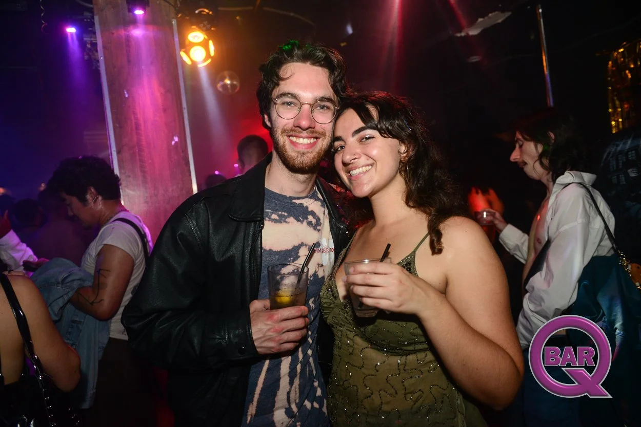 Two young adults, a man with glasses and a beard wearing a black jacket and a woman with curly dark hair in a green top, smiling and holding drinks, enjoying a night at a bar or club with colorful lighting and other patrons in the background.