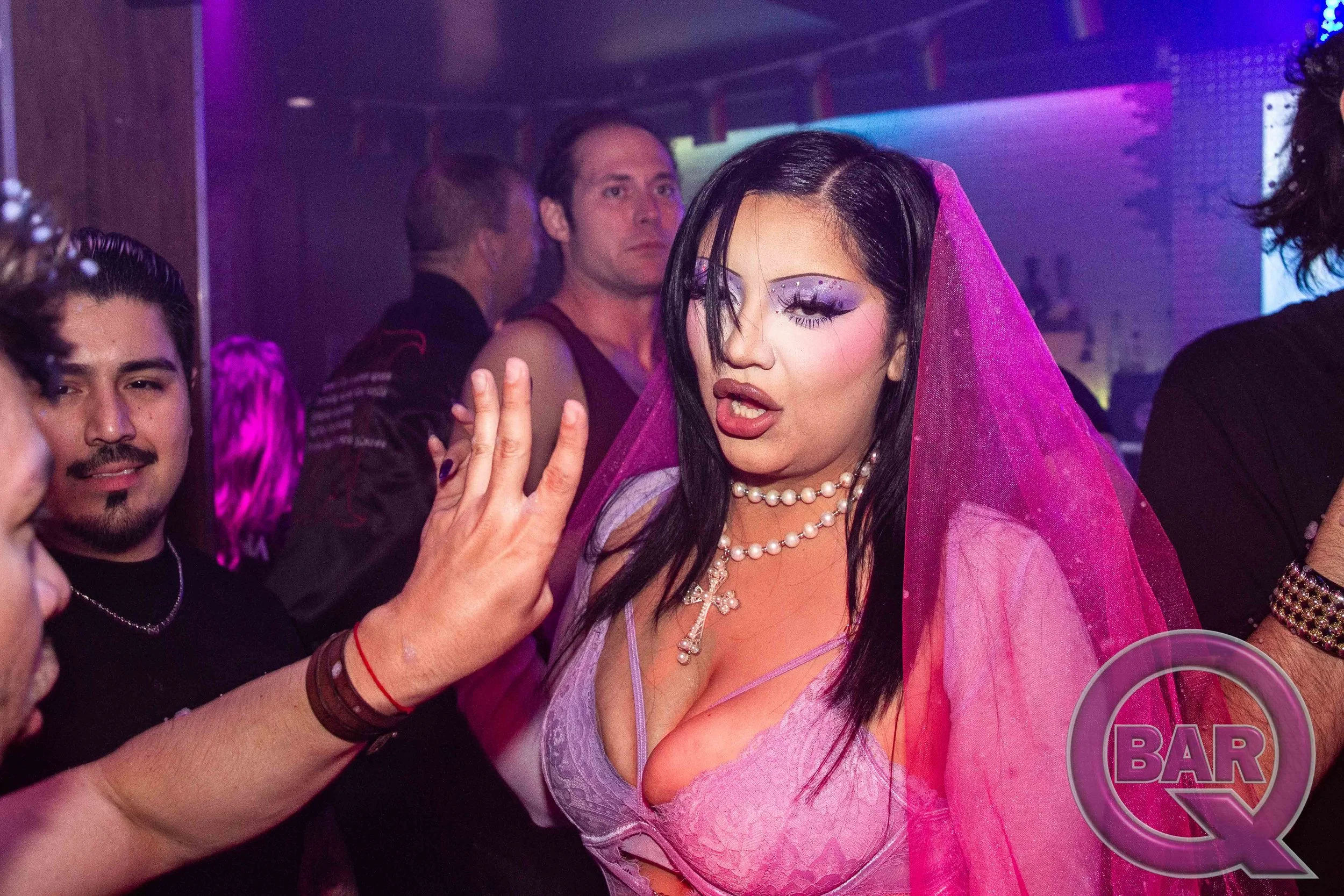 A woman with dark hair and elaborate makeup, wearing a pink lace outfit and pearl necklace, at a nightclub or bar. She is making a gesture with her hand, possibly part of a conversation or dance.