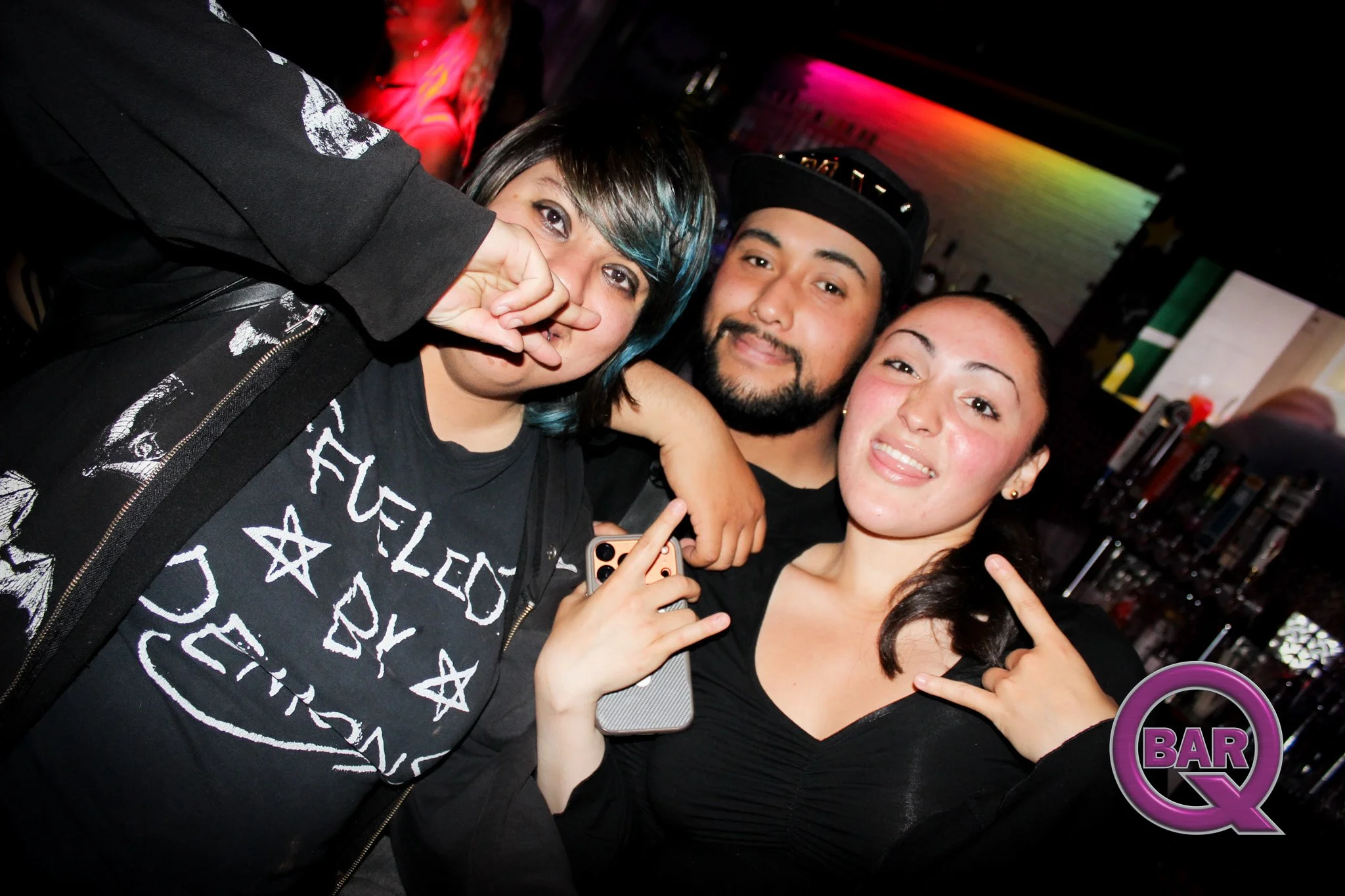 Group of four friends smiling and making peace signs at a bar or nightclub with colorful lighting.