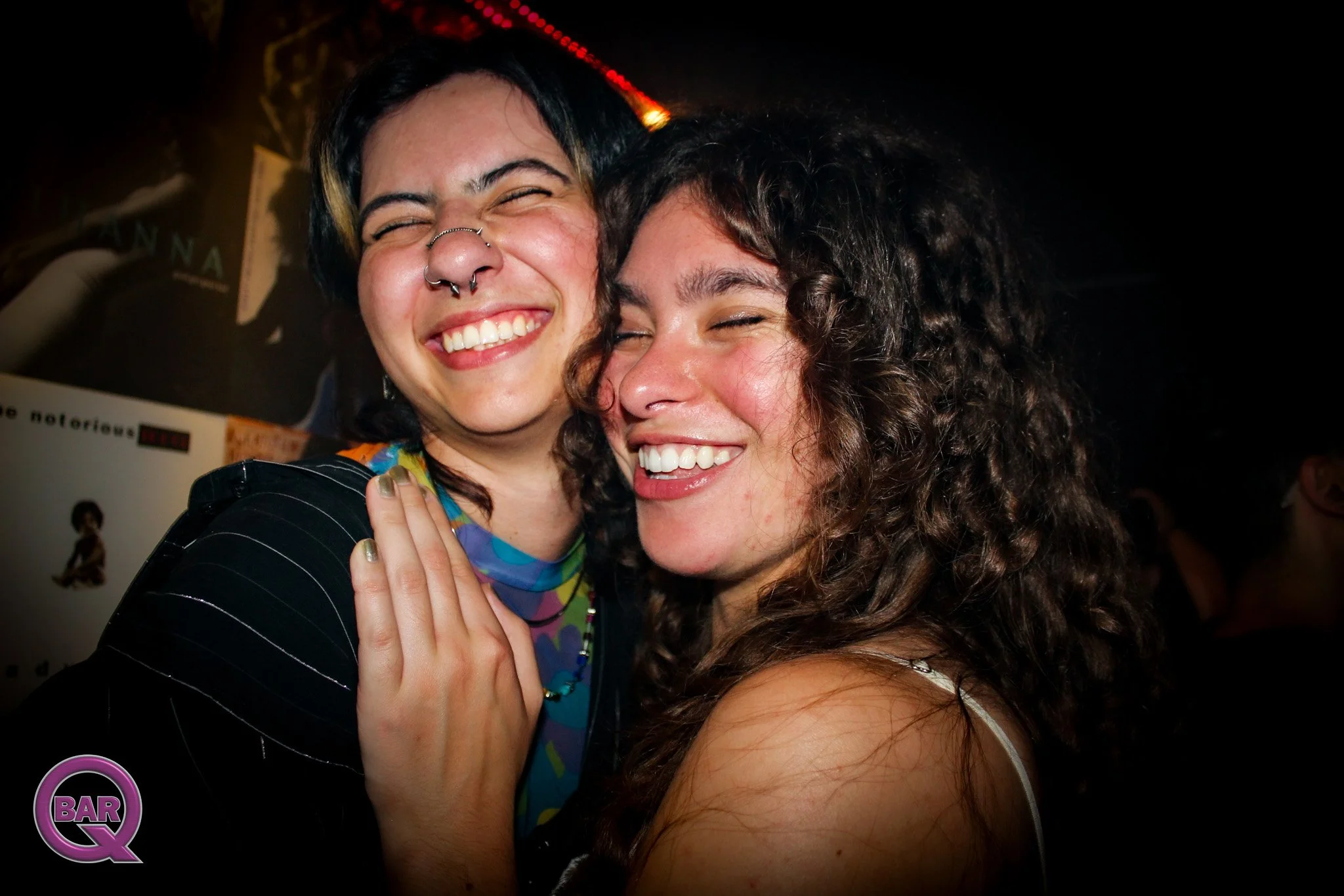 Two women with big smiles, hugging each other, at a night event, with a dark background and some red light and a DJ booth visible.