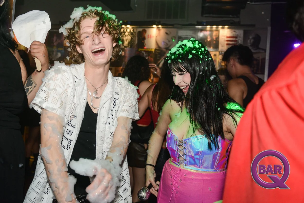 Two people, a young man and woman, are laughing and having fun at a party or club. The young man has curly hair, is wearing a white lace shirt, and has foam on his arms and head. The woman has long black hair, is wearing an iridescent top and pink sk