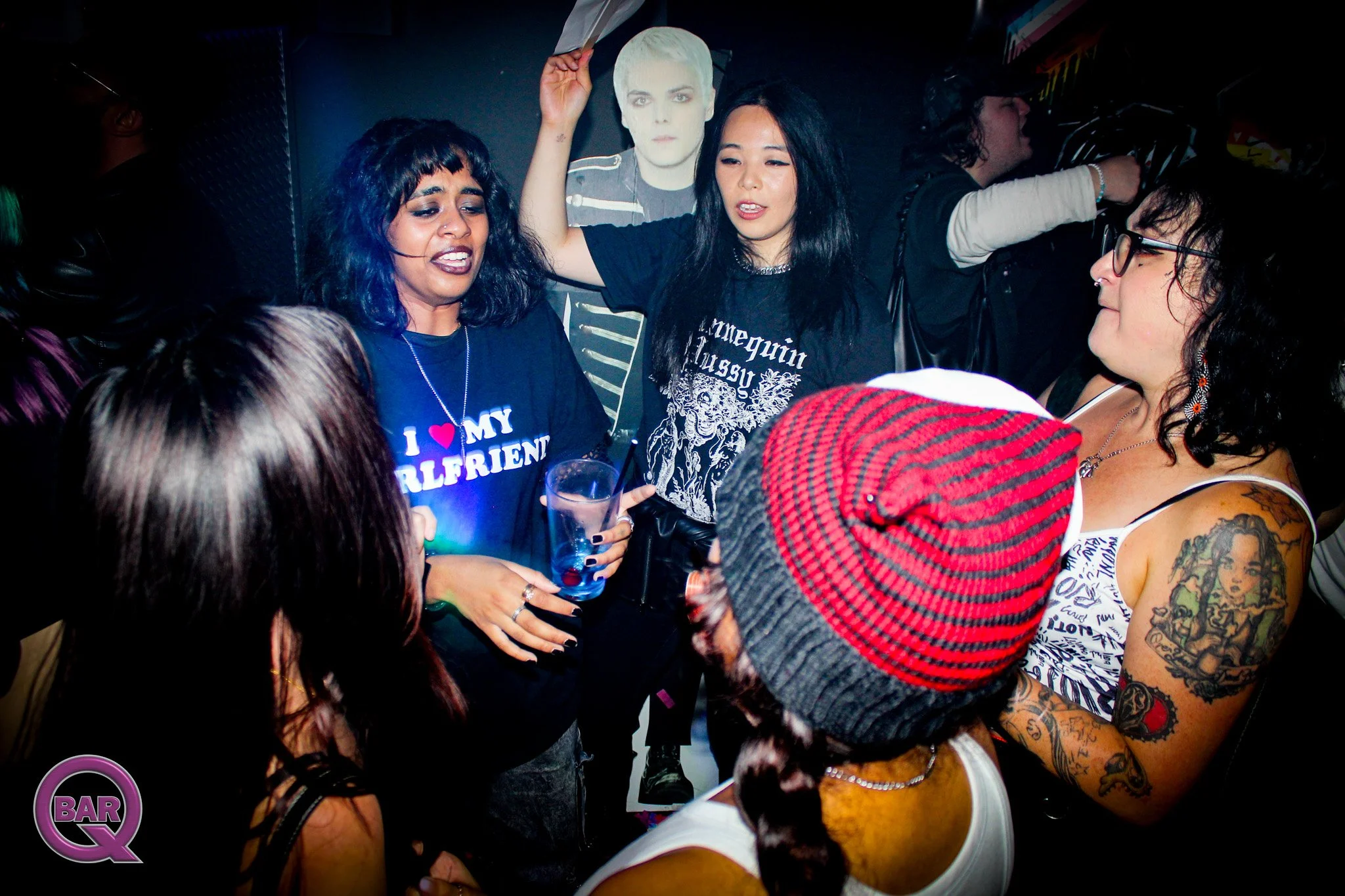 Group of young women and men socializing at a bar or club, with some holding drinks and tattoos visible on one woman's arm.