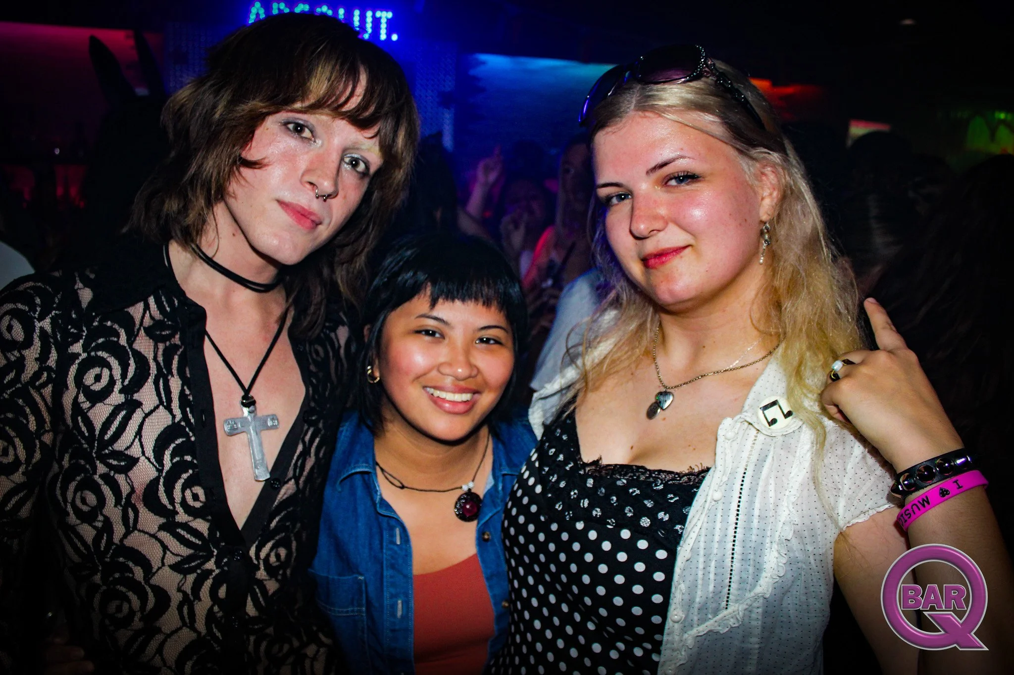 Four women posing together at a nightclub or bar, smiling and wearing casual and stylish clothing, with colorful lighting and a sign that reads 'bar' in the background.