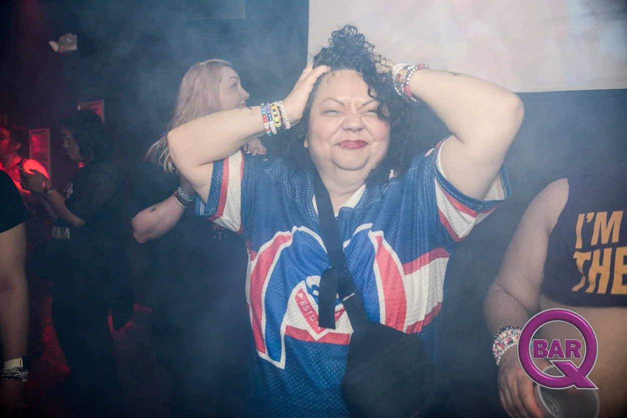 Woman in a sports jersey with her hands on her head, smiling with eyes closed at a bar or club, with a smoky atmosphere and other people in the background.