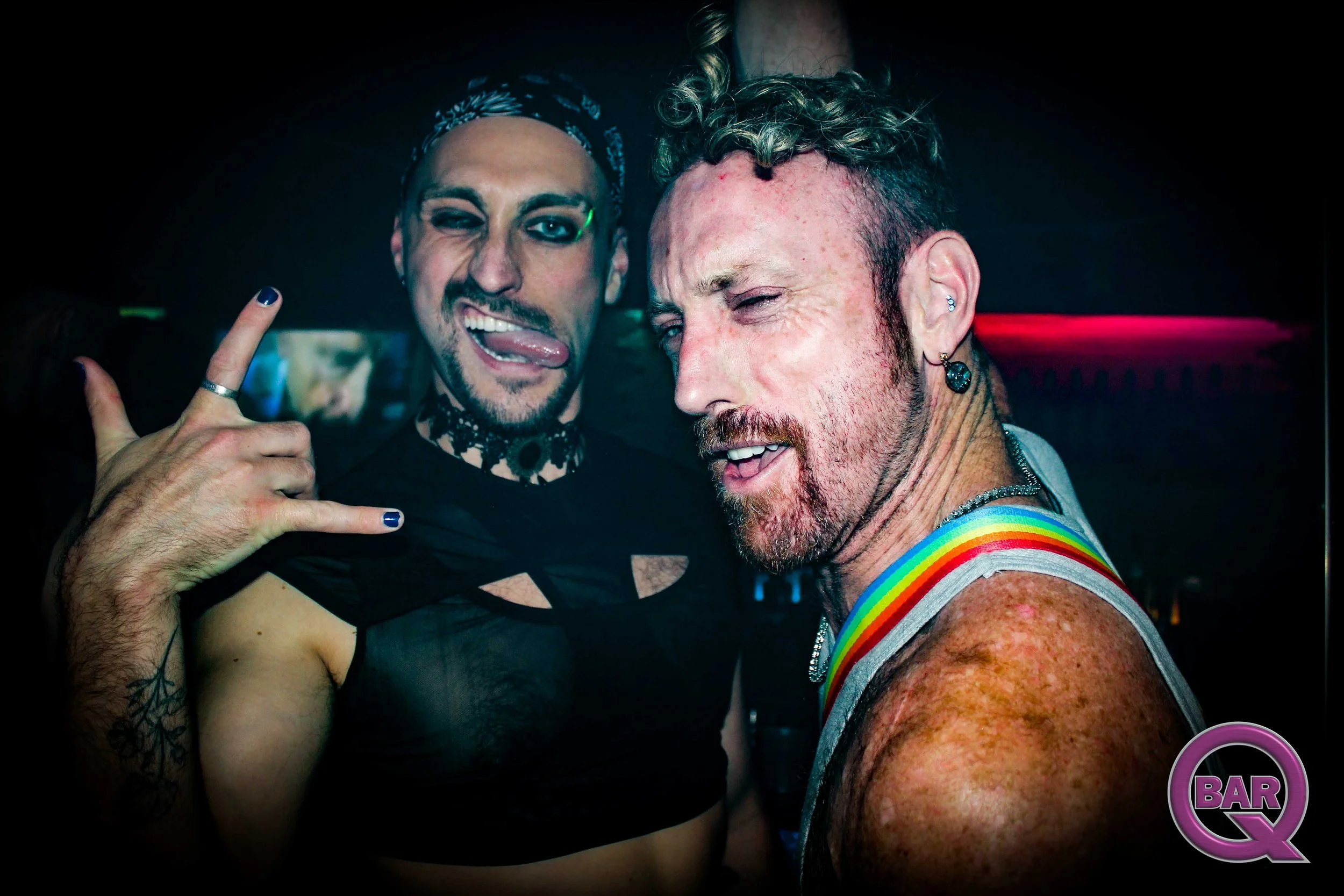 Two men having fun at a party or club; one is winking and making a peace sign, while the other is smiling with colorful makeup and rainbow suspenders, amidst a dark, lively environment.