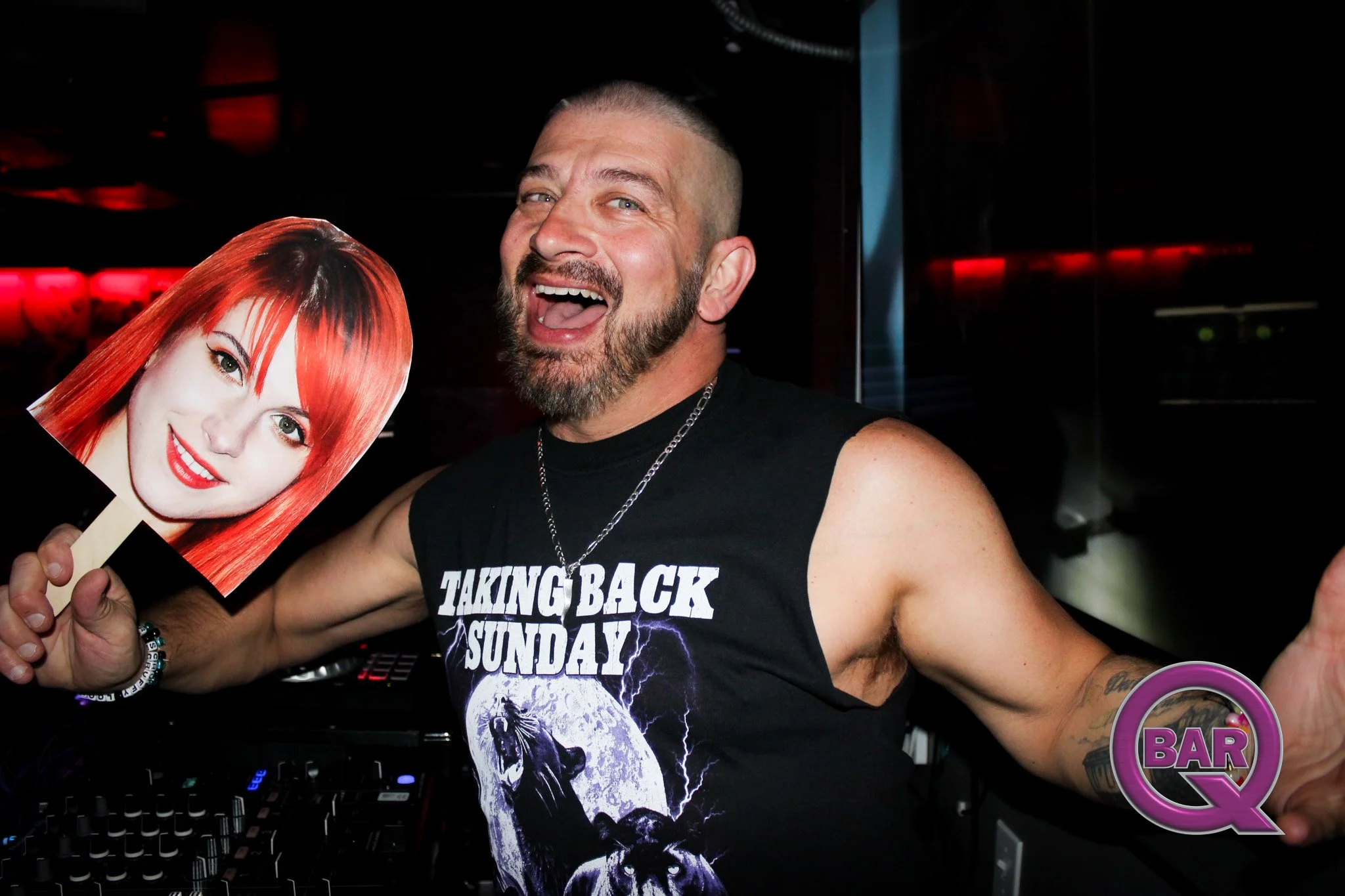 A man with a beard and short hair smiling and holding a picture of a woman with red hair and red lipstick in front of a dark background with red lighting.