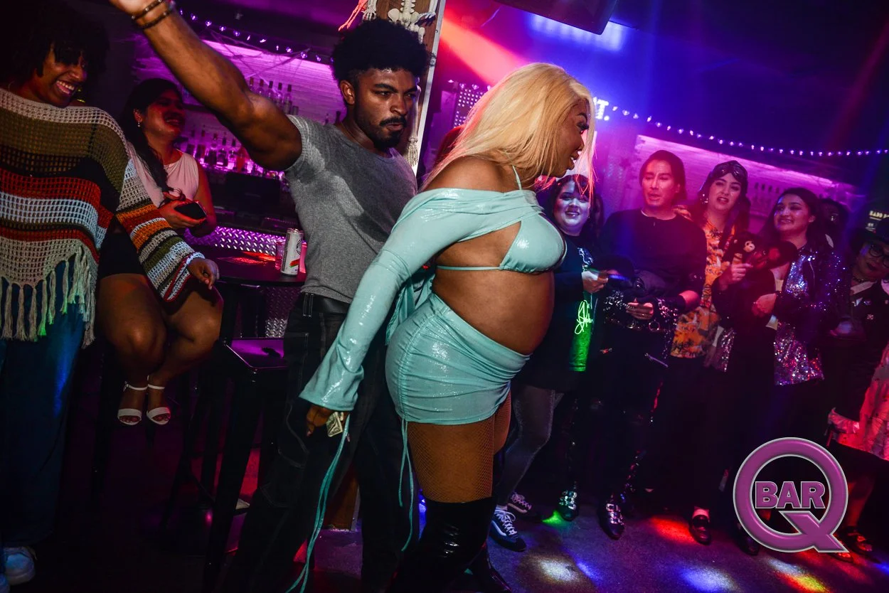 A group of people dancing and socializing in a nightclub with colorful lighting, featuring a woman in a shiny turquoise outfit in the center.