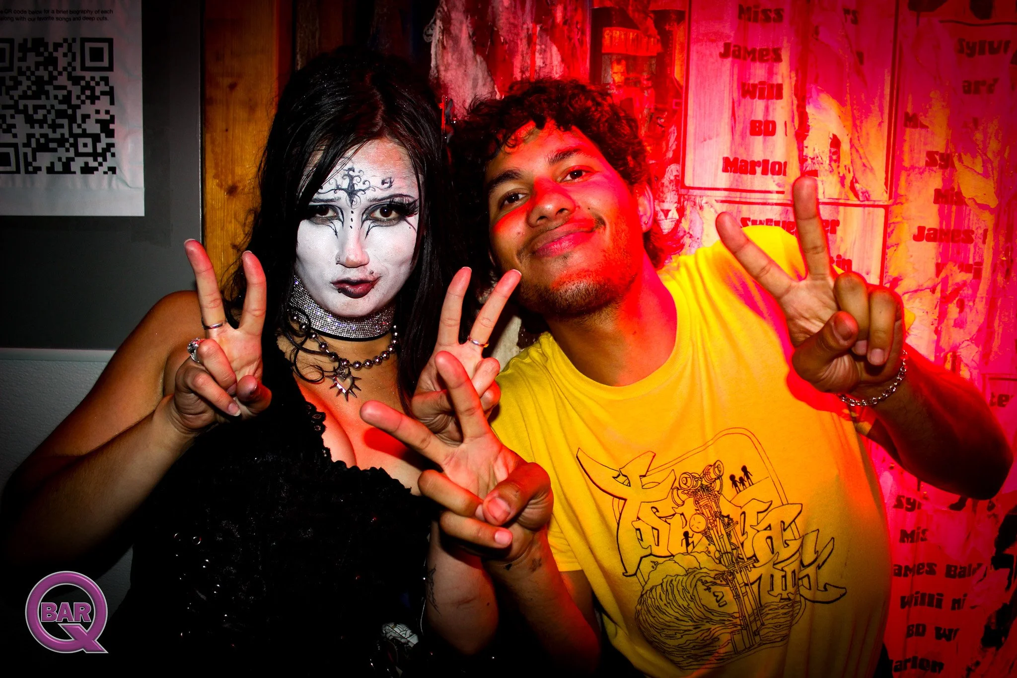 Two people posing with peace signs at a bar or nightclub. One person has elaborate face makeup and dark hair, wearing black attire and jewelry. The other person has curly hair, light brown skin, and is wearing a yellow T-shirt with a graphic design. 