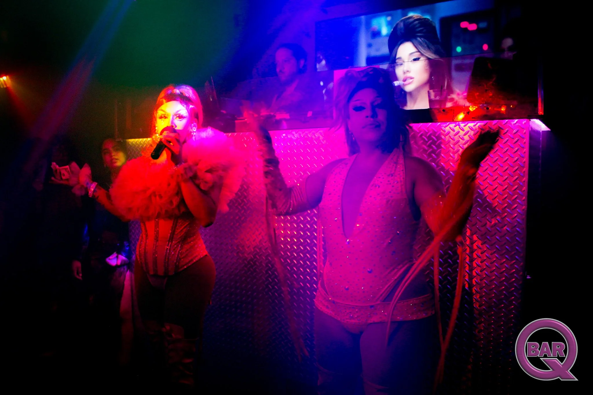 A drag performer dressed in a vibrant pink outfit with feathers, holding a microphone and performing on stage in a nightclub. The background features reflections of the performer and a woman with dark hair is seen on a mirror. The scene is illuminate