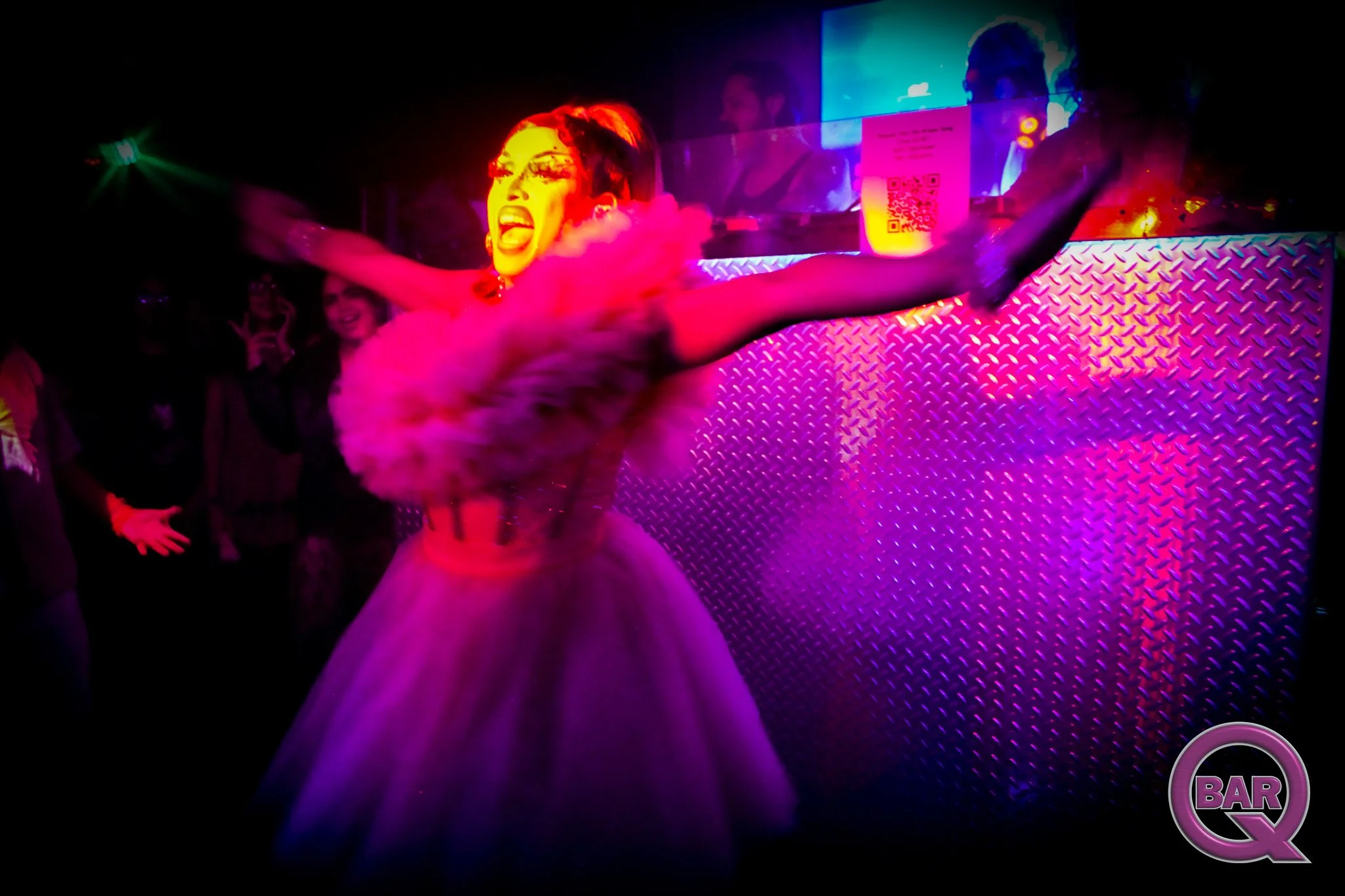A performer with red hair and makeup, wearing a colorful outfit, dancing in a nightclub with colorful lights and a DJ in the background.
