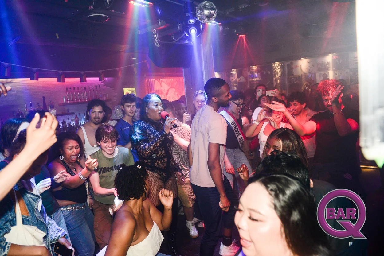 A crowd dancing and singing in a nightclub with colorful lights, a woman with a microphone in the center, and a disco ball overhead.