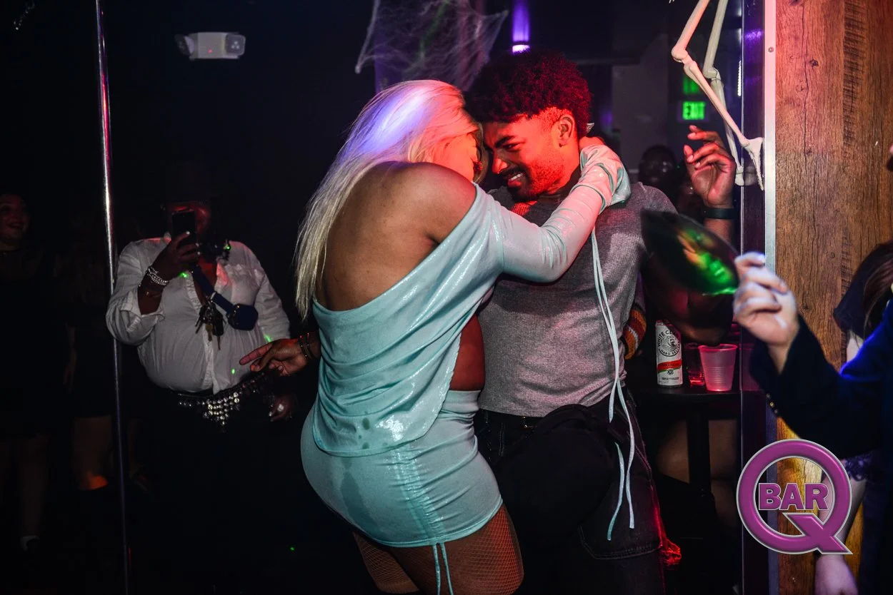 A woman and a man are dancing closely and smiling at each other in a dimly lit nightclub with neon lights. The woman has platinum blonde hair and is wearing a shiny, light-colored dress with long sleeves. The man has curly dark hair and a beard, wear