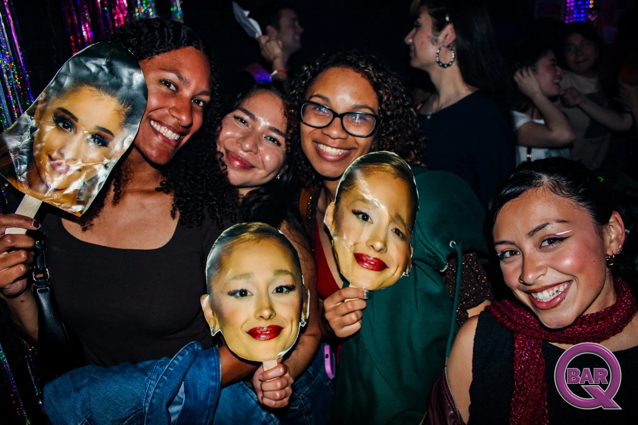 Group of diverse women at a party, holding masks with a woman's face and a 'BAR' Q in a colorful environment.