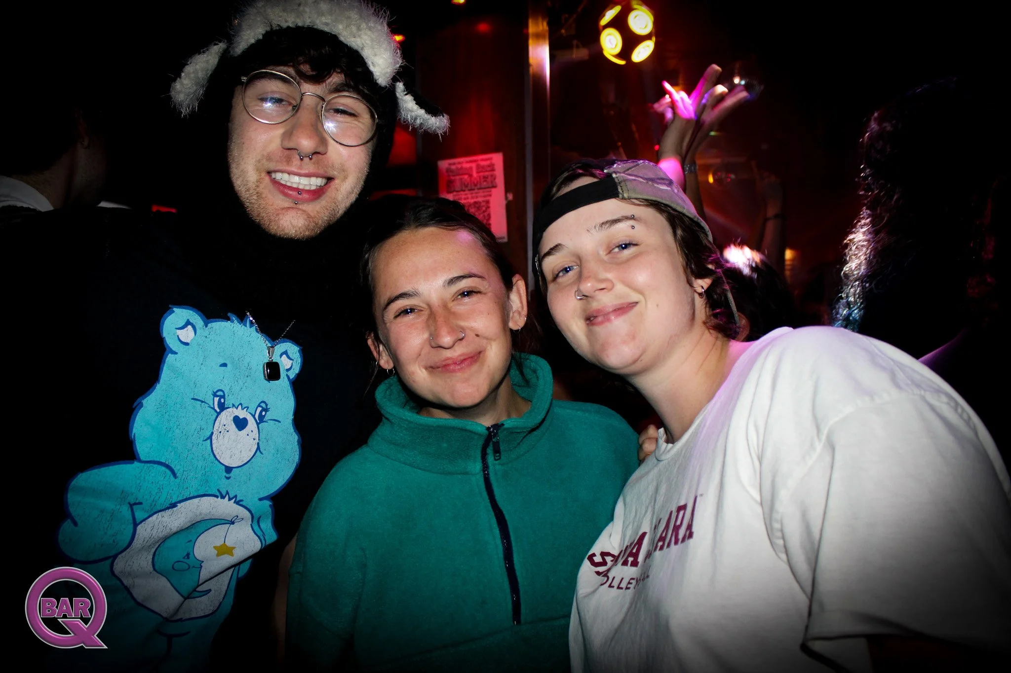 Three people smiling at the camera at a party or nightclub. One person is wearing glasses and a festive hat, another is wearing a teal hoodie, and the third is wearing a white shirt and a backwards cap. The background has dark lighting with some brig