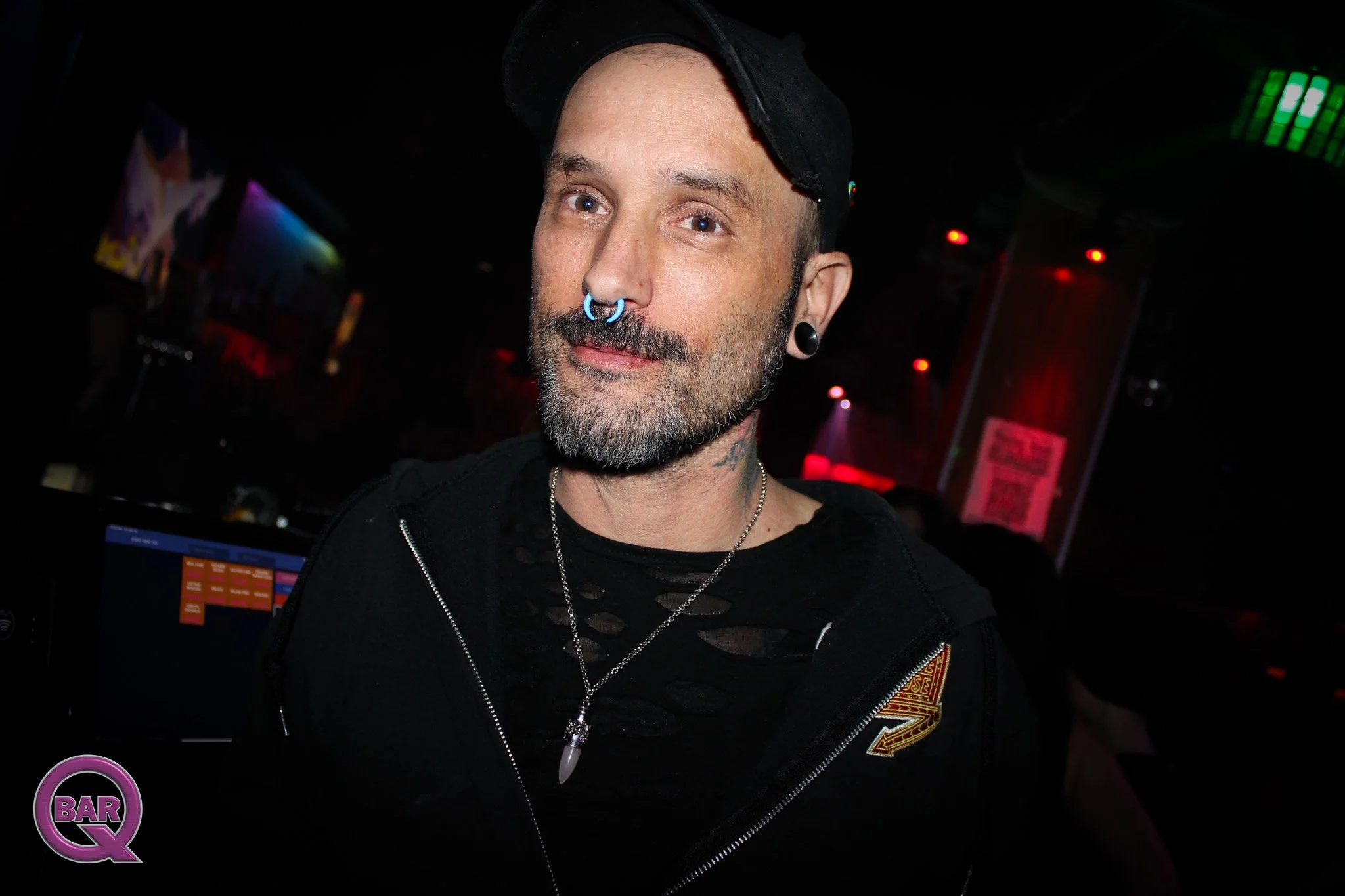 A man with a beard and tattoos on his neck, wearing a black baseball cap backwards, black earrings, a septum piercing, a black ripped shirt, and necklaces, standing in a nightclub with colorful lights.